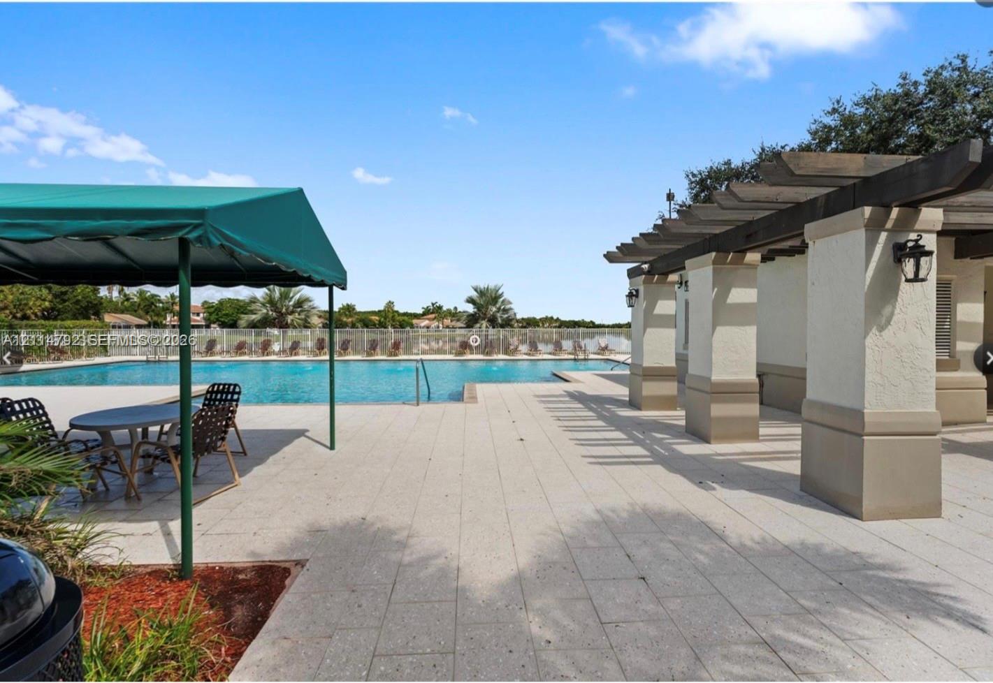 Outdoor pool area featuring a stone-paver patio, wood-beam pergola, green canvas awning, and metal perimeter fencing