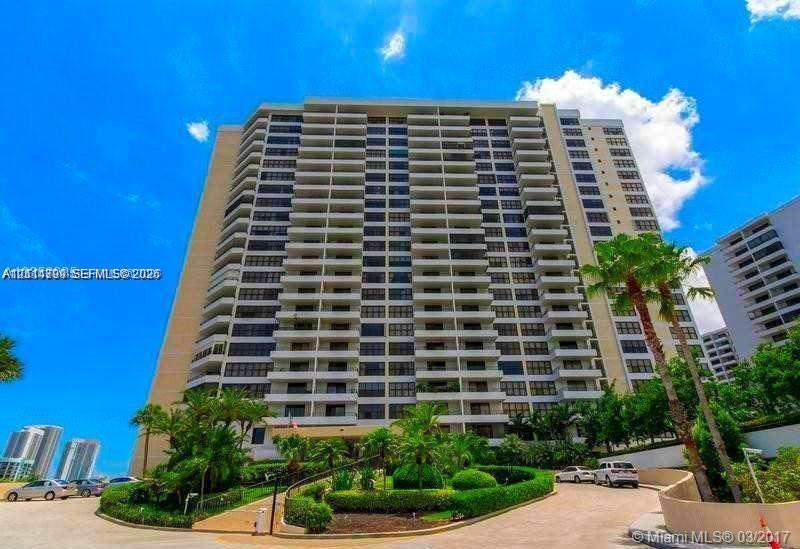 High-rise residential building featuring expansive private balconies, a paved arrival drive, and manicured landscaping with mature palm trees