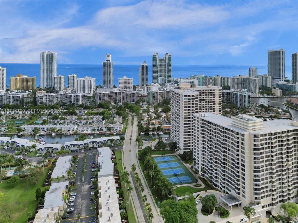 Aerial perspective featuring coastal urban surroundings, a multi-story high-rise complex, tennis courts, and mature landscaping