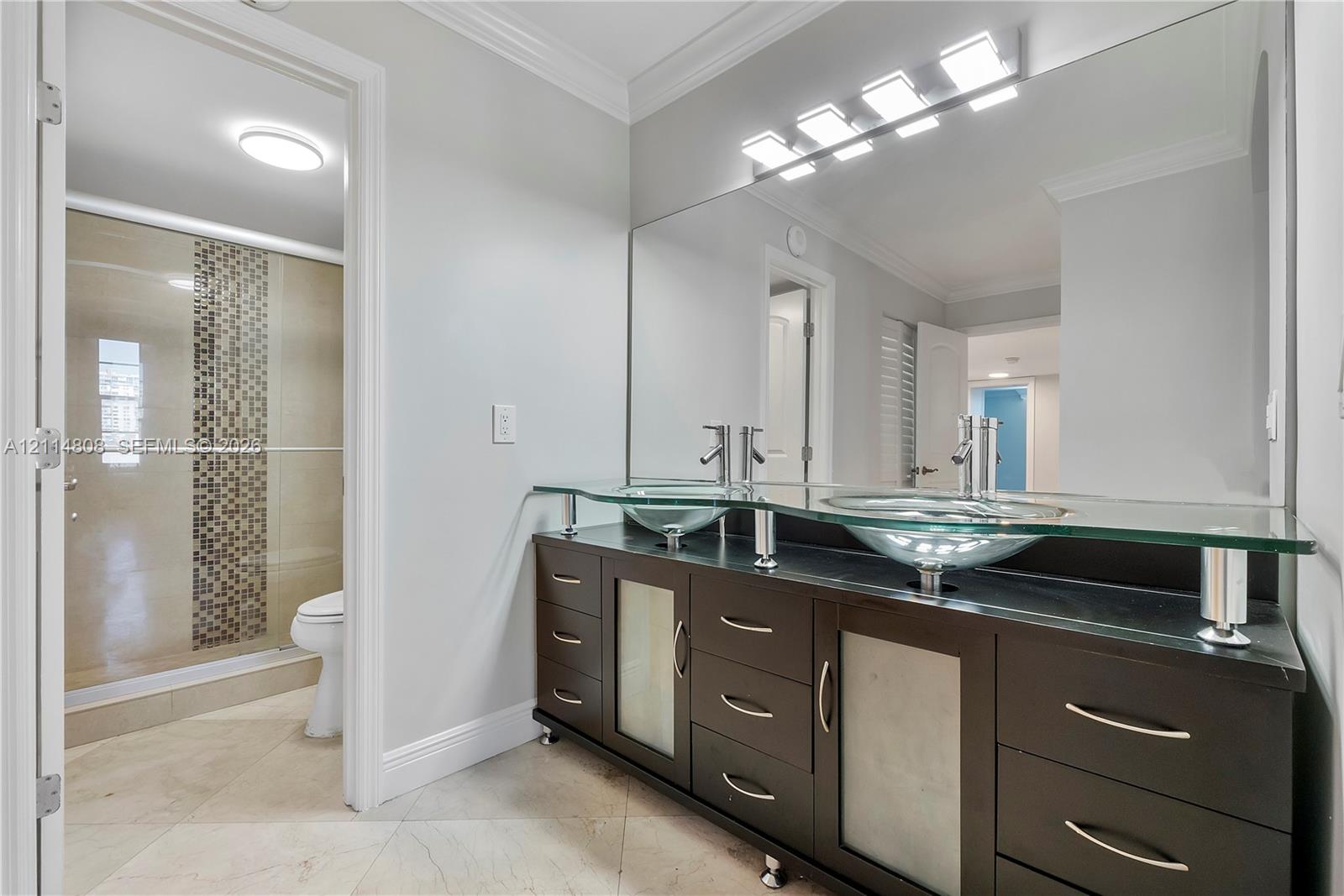 Double vanity with vessel sinks, glass countertop, dark wood-finish cabinetry, full-width mirror, and walk-in shower with mosaic tile accent