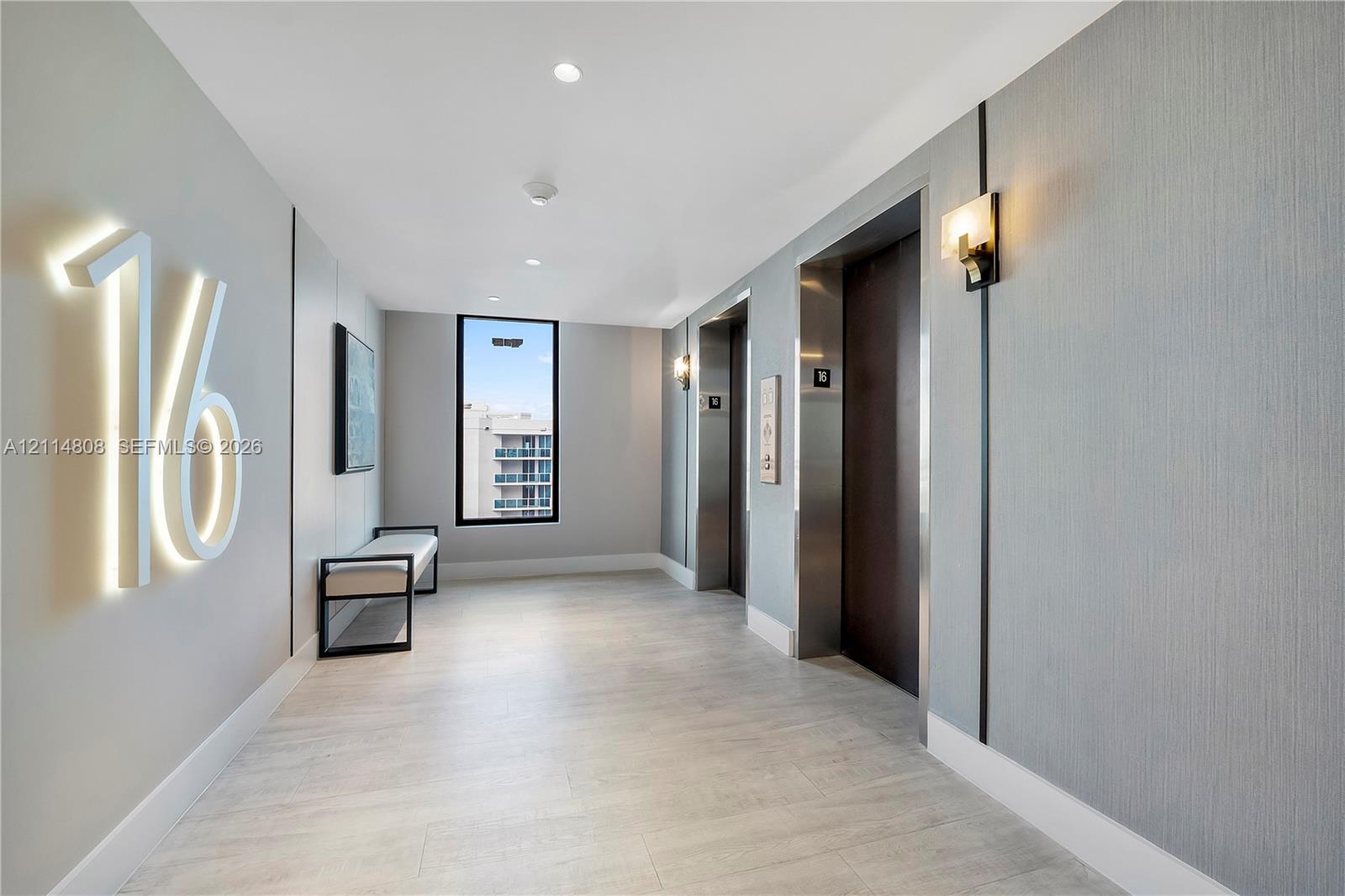 Elevator lobby featuring illuminated wall signage, recessed ceiling lighting, textured wall coverings, dual elevator access with metallic finishes, and wood-finish flooring