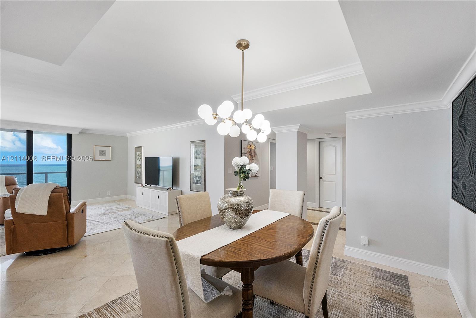 Open-concept interior featuring light-toned stone tile flooring, tray ceilings with crown molding, a multi-globe pendant light fixture, and floor-to-ceiling windows with ocean scenery
