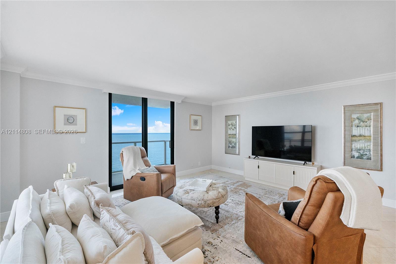 Living area featuring light-toned tile flooring, crown molding, and floor-to-ceiling glass sliding doors leading to an exterior balcony with water scenery