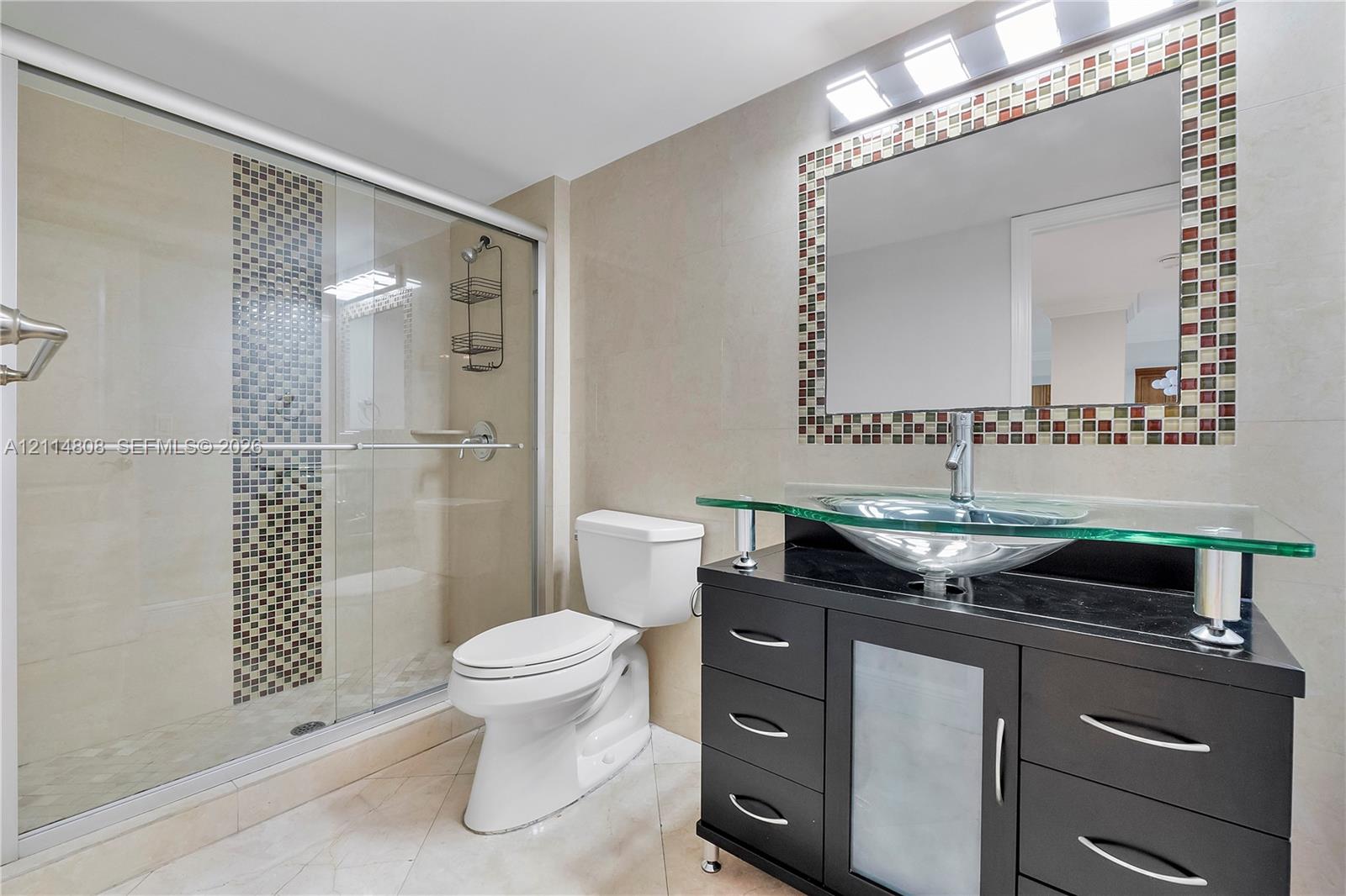 Modern vanity featuring a black finish, glass countertop, and vessel sink, paired with a glass-enclosed shower with mosaic tile accents, matching mosaic tile framed mirror, and light-toned tile flooring
