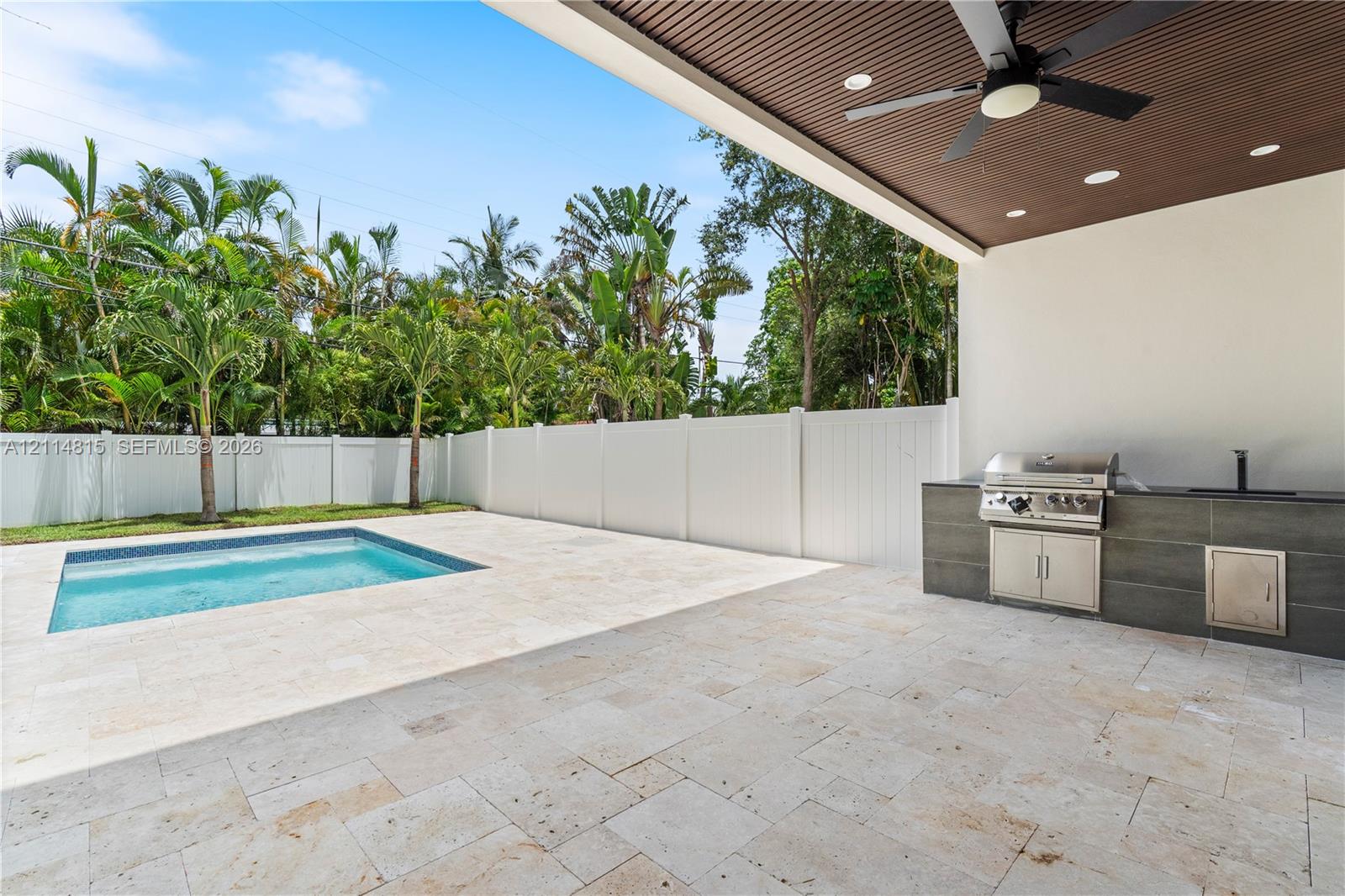 Covered outdoor kitchen with stainless steel grill, stone-finish cabinetry, built-in ceiling fan, recessed lighting, and travertine patio surrounding a swimming pool enclosed by vinyl fencing