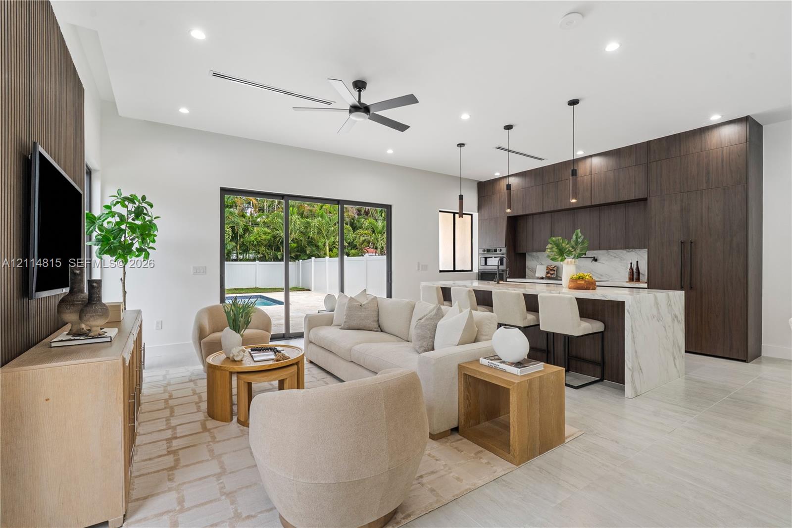 Open-concept interior featuring wood-finish cabinetry, a waterfall-edge kitchen island, stone-surfaced countertops, sliding glass doors, and light-toned tile flooring Open-concept interior featuring wood-finish cabinetry, a waterfall-edge kitchen island, stone-surfaced countertops, sliding glass doors, and light-toned tile flooring