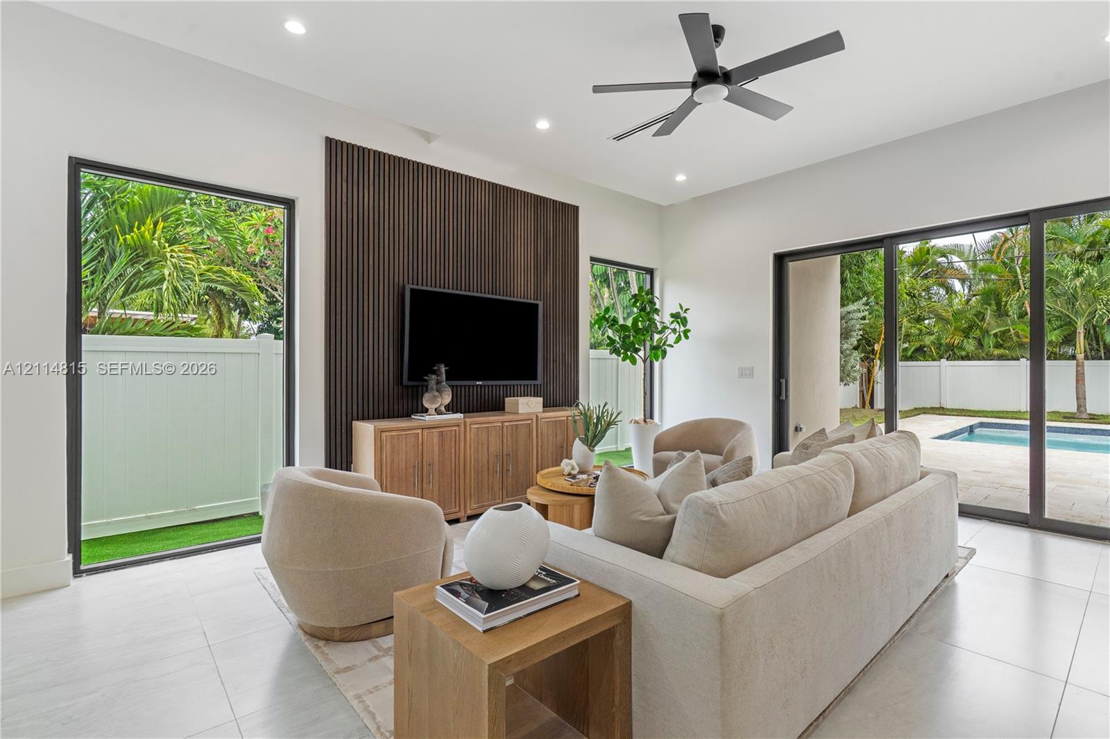 Living area featuring large-format tile flooring, a dark-toned vertical slat accent wall, recessed lighting, a modern ceiling fan, and sliding glass doors with direct access to the exterior pool area Living area featuring large-format tile flooring, a dark-toned vertical slat accent wall, recessed lighting, a modern ceiling fan, and sliding glass doors with direct access to the exterior pool area