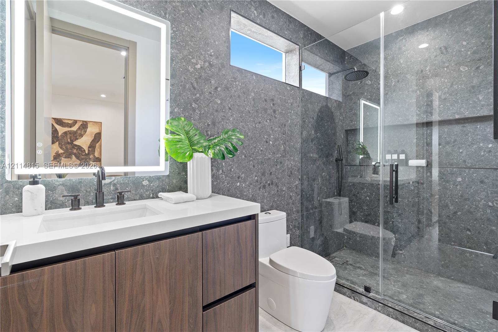 Bathroom featuring a wood-finish vanity with a quartz countertop, matte black fixtures, an integrated LED-backlit mirror, and a walk-in shower with floor-to-ceiling stone-style tile and built-in bench