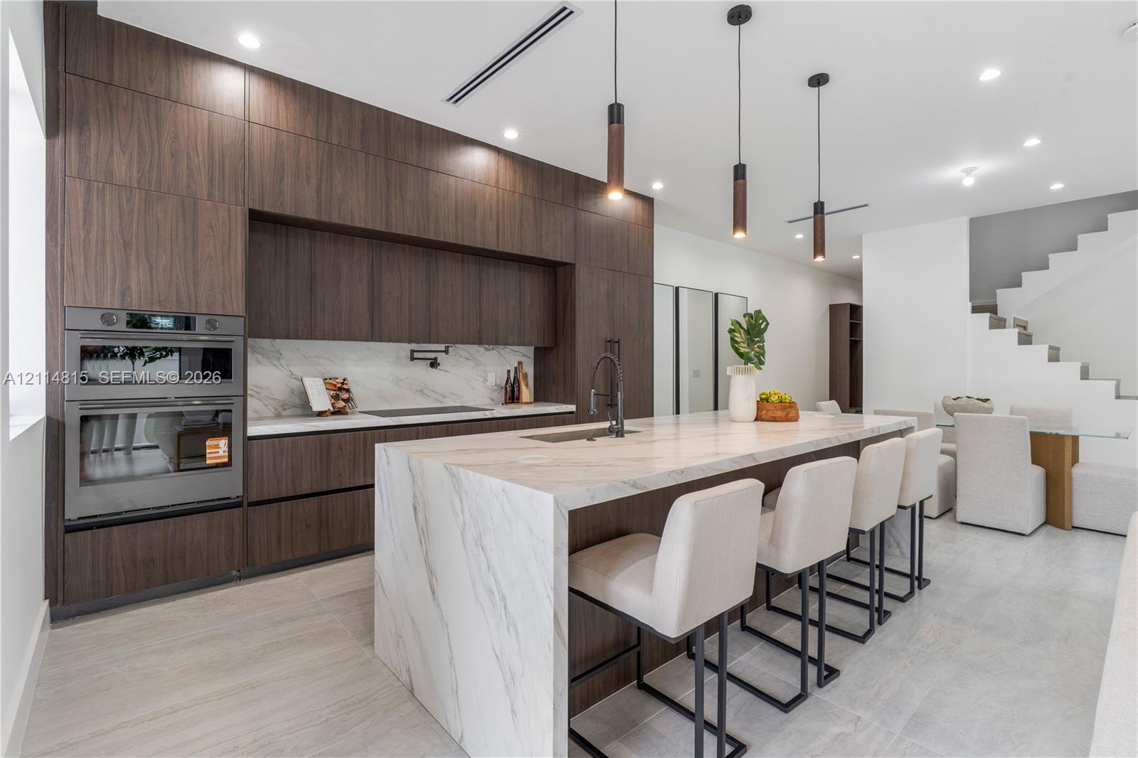 Kitchen featuring dark wood-finish cabinetry, a waterfall-edge stone island with an integrated sink, built-in double wall ovens, a pot filler faucet, and pendant lighting Kitchen featuring dark wood-finish cabinetry, a waterfall-edge stone island with an integrated sink, built-in double wall ovens, a pot filler faucet, and pendant lighting