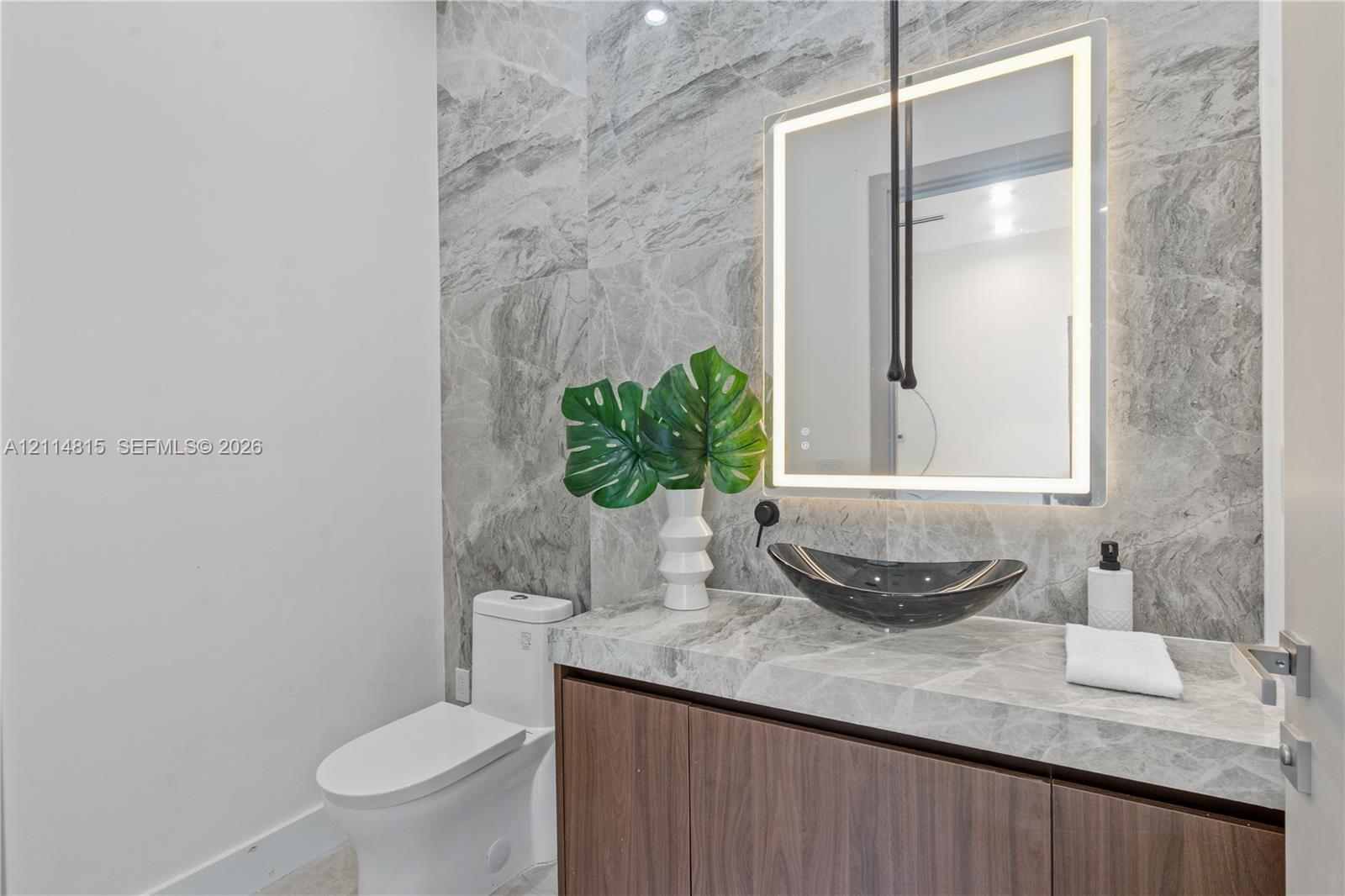 Modern vanity with wood-finish cabinetry, stone countertop, vessel sink, and wall-mounted faucet, complemented by full-height stone wall tiling and a rectangular backlit LED mirror Modern vanity with wood-finish cabinetry, stone countertop, vessel sink, and wall-mounted faucet, complemented by full-height stone wall tiling and a rectangular backlit LED mirror