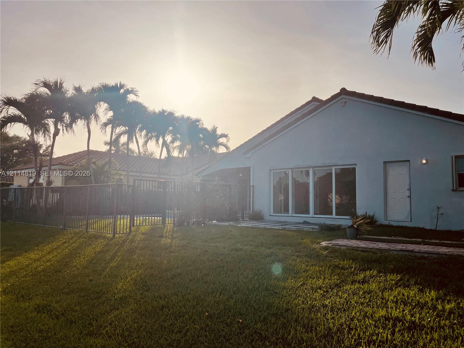 Exterior rear elevation featuring a concrete patio, metal perimeter fencing, tiled roof, and mature palm trees