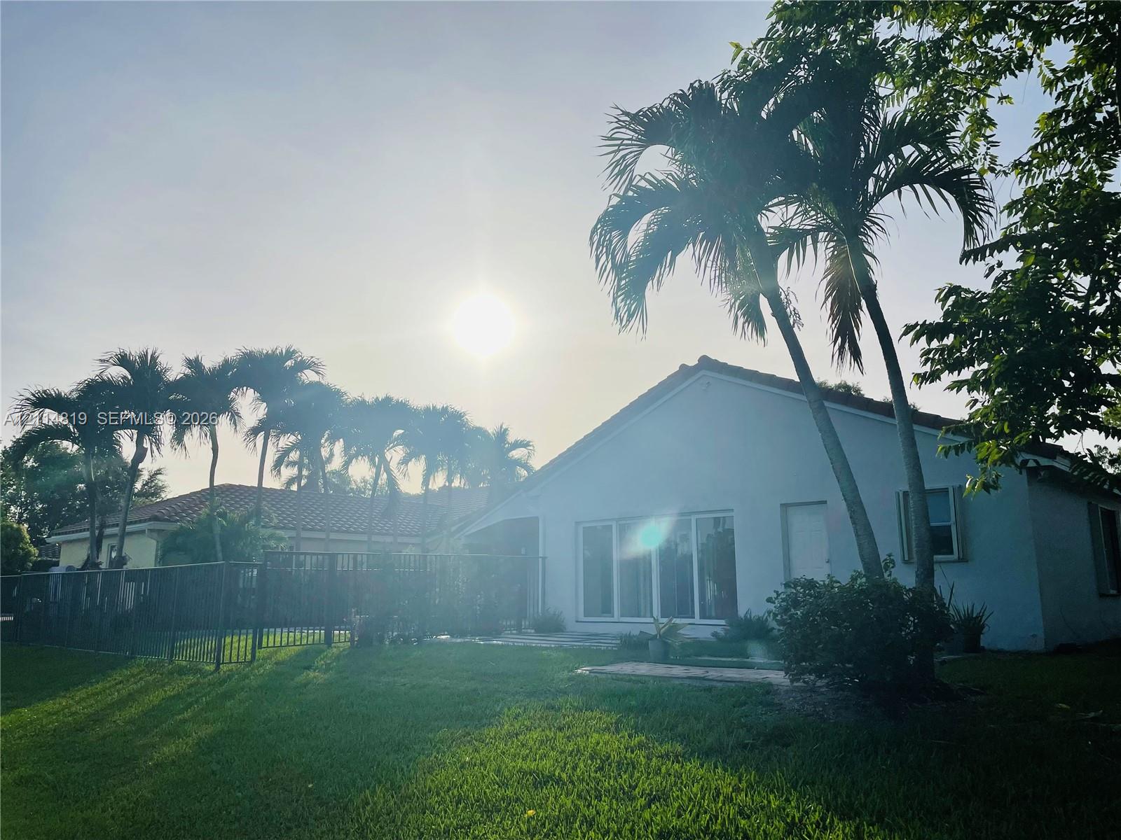 Exterior elevation featuring a stucco facade, tiled roof, glass sliding doors, and mature palm trees within a fenced yard