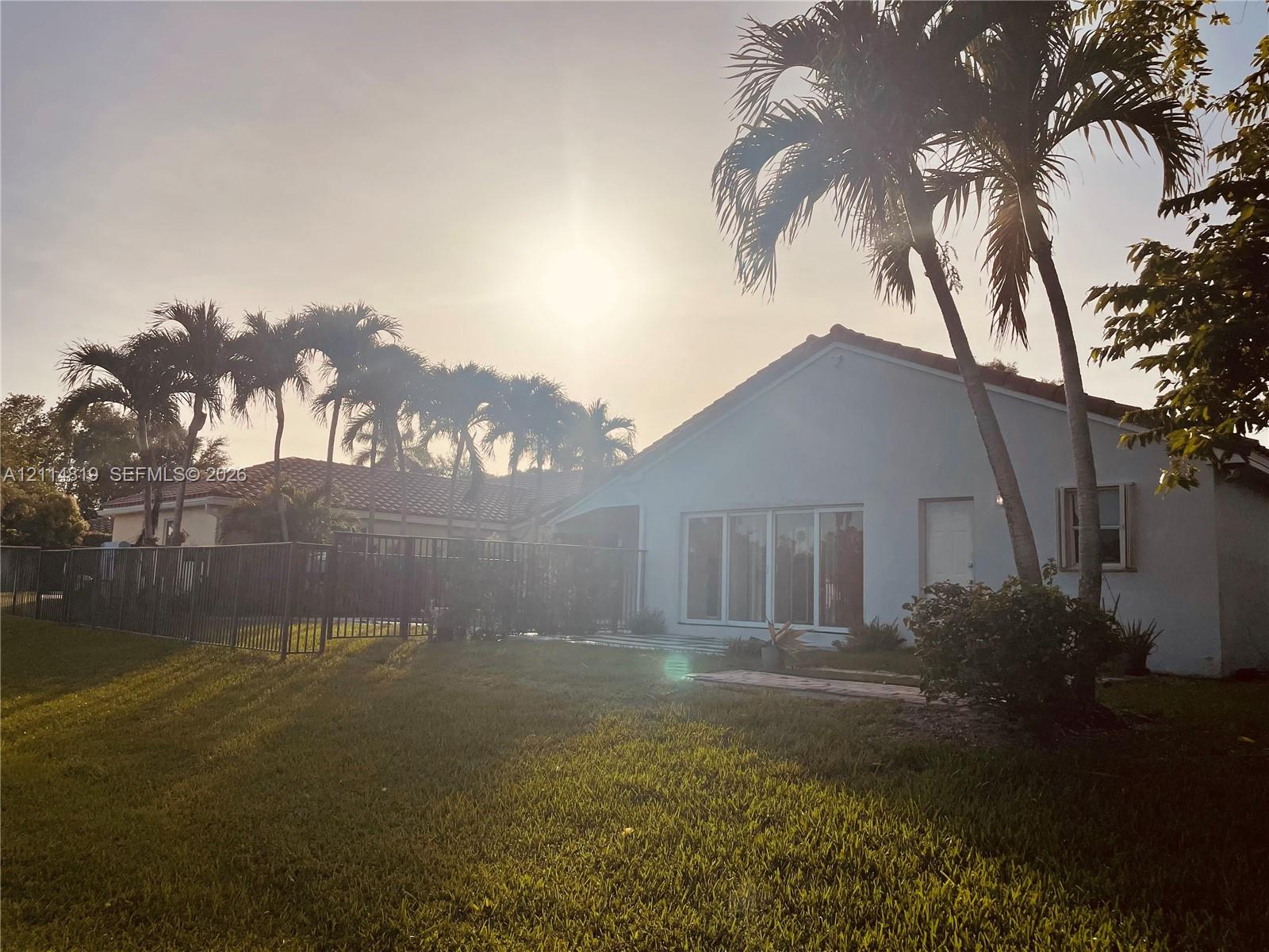 Exterior rear elevation featuring a stucco finish, metal-framed glass sliding doors, a tiled roof, mature palm trees, and a fenced yard