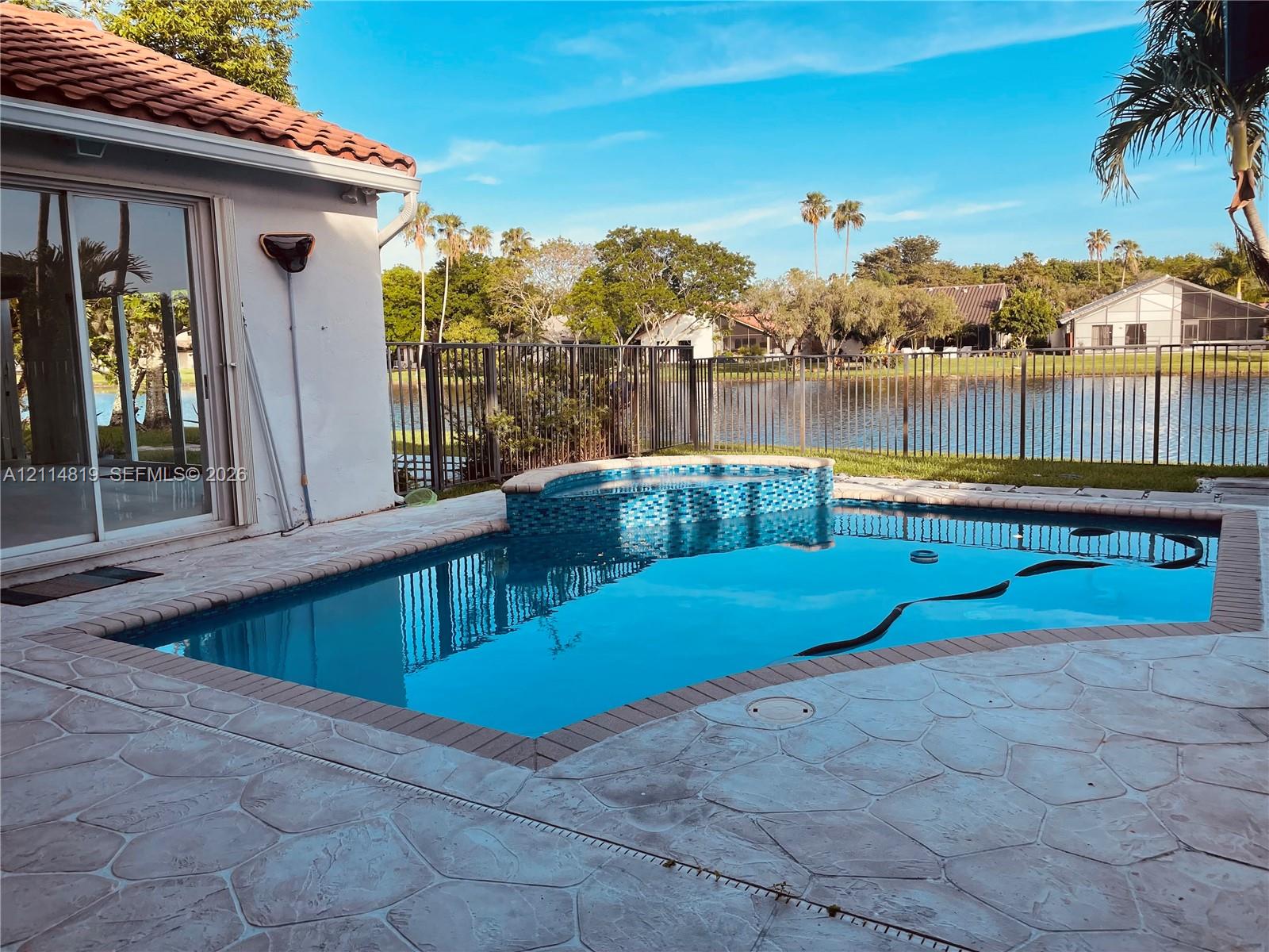 In-ground swimming pool with an attached raised spillover spa, tiled water line, concrete paver decking, and metal perimeter fencing overlooking a pond