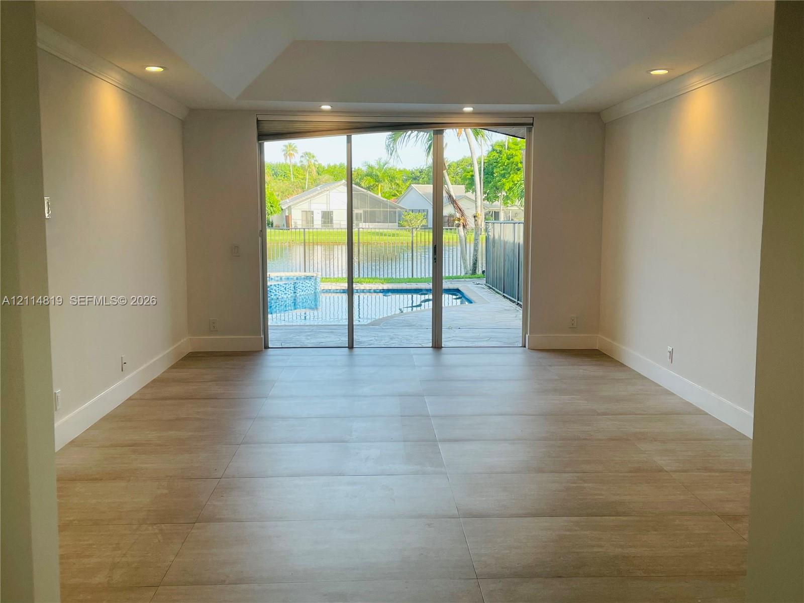 Tray ceiling, large-format tile flooring, recessed lighting, and expansive sliding glass doors opening to a private pool and water-front lot