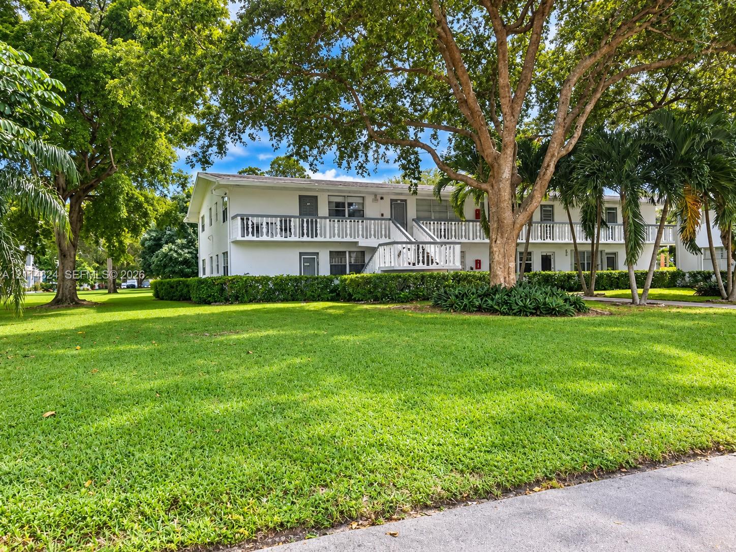 Two-story exterior featuring white stucco siding, expansive wrap-around balconies with railings, mature shade trees, and manicured landscaping
