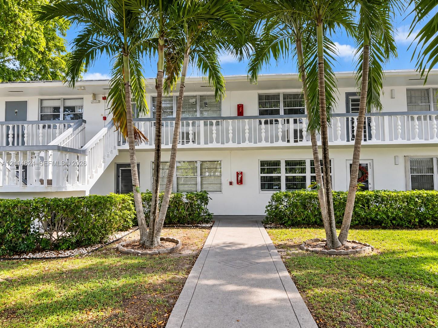 Multi-story exterior featuring white stucco siding, a second-level balcony with white spindle railing, concrete walkway, and landscaped palm trees Multi-story exterior featuring white stucco siding, a second-level balcony with white spindle railing, concrete walkway, and landscaped palm trees