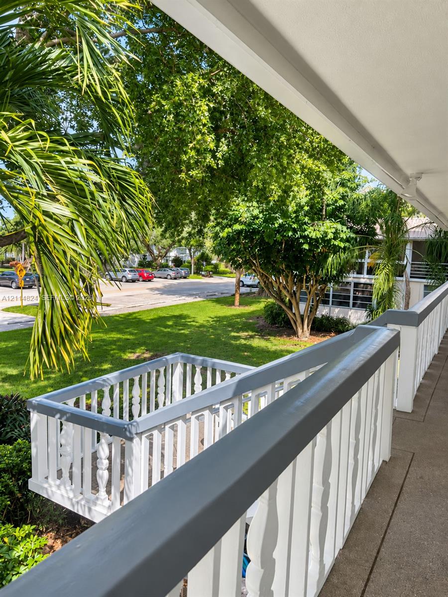 Covered balcony with a gray-painted wood railing, featuring balusters and a concrete floor surface overlooking a landscaped lawn with mature trees and palms