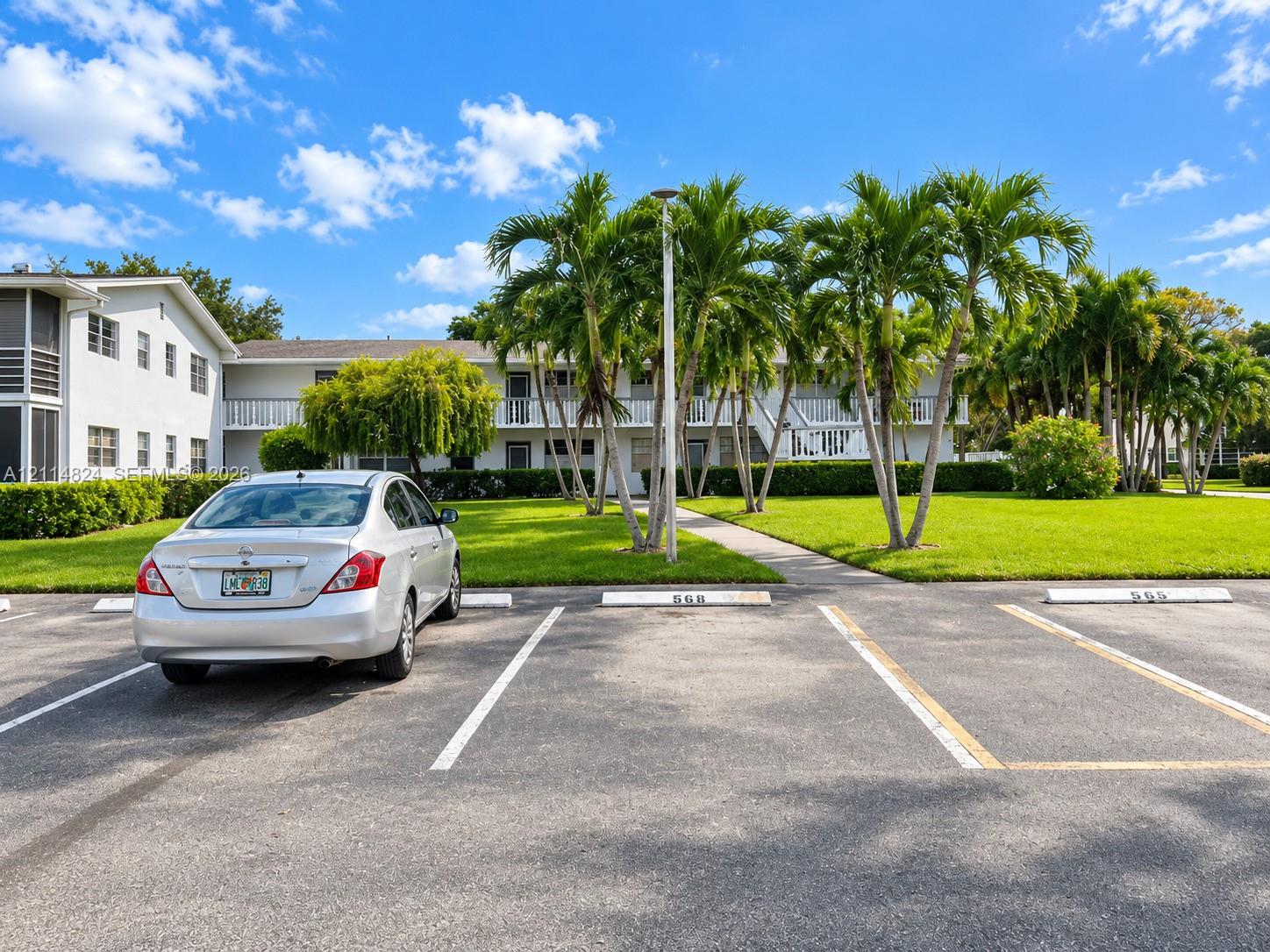 Two-story exterior featuring a light-colored facade, private balconies with railings, mature palm trees, and an asphalt parking area with designated numbered spaces