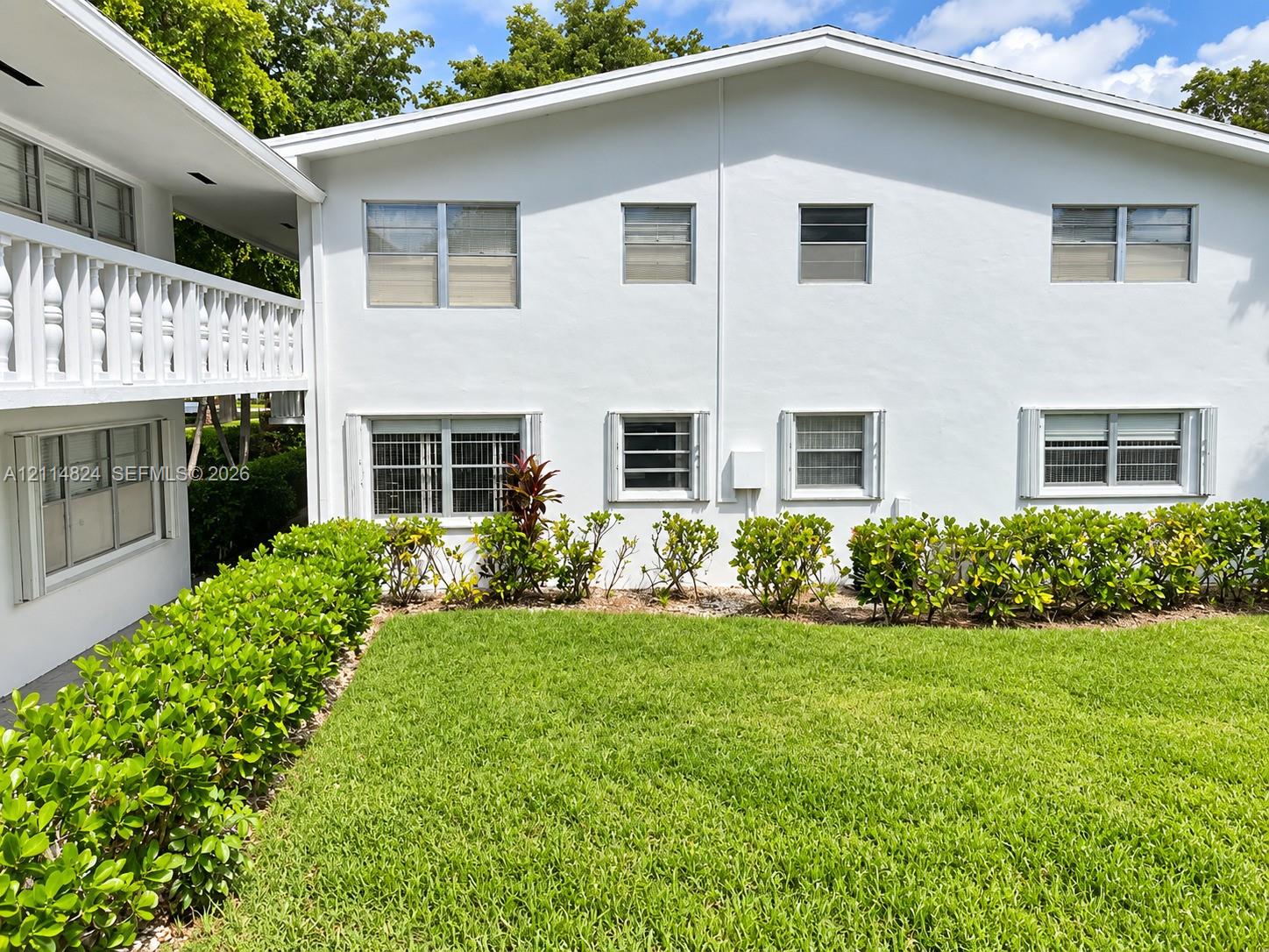 Multi-story exterior featuring white stucco finish, second-level balcony with decorative railings, manicured lawn, and established hedge landscaping
