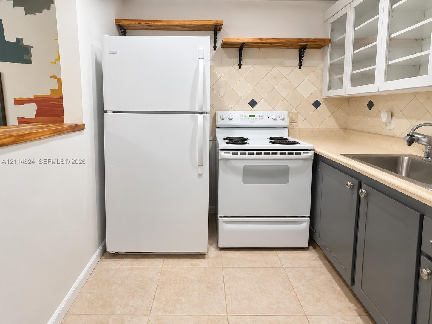 Kitchen featuring charcoal shaker-style cabinetry, white appliances, wall-mounted wood shelving, diamond-patterned tile backsplash with inset accents, and tile flooring