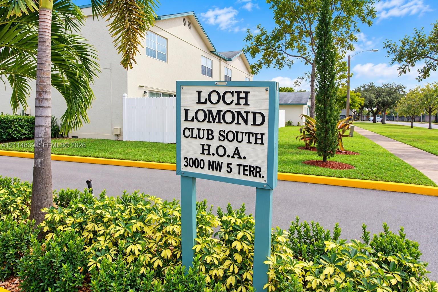 Exterior featuring a light-toned stucco facade, white vinyl fencing, paved asphalt pathways, and mature palm and cypress landscaping