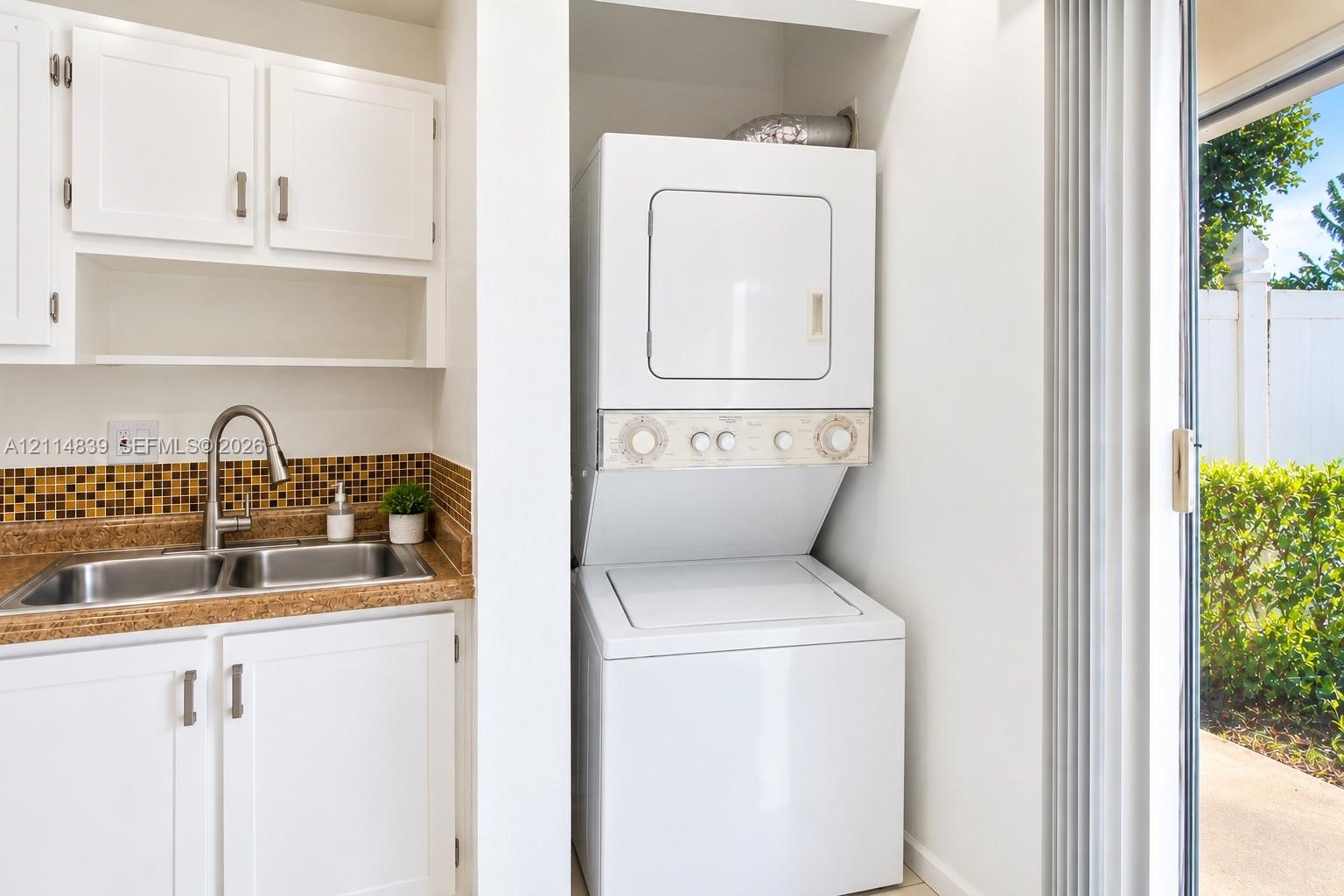 Laundry area featuring white cabinetry, a double-basin stainless steel sink, a mosaic tile backsplash, and a stacked washer-dryer unit