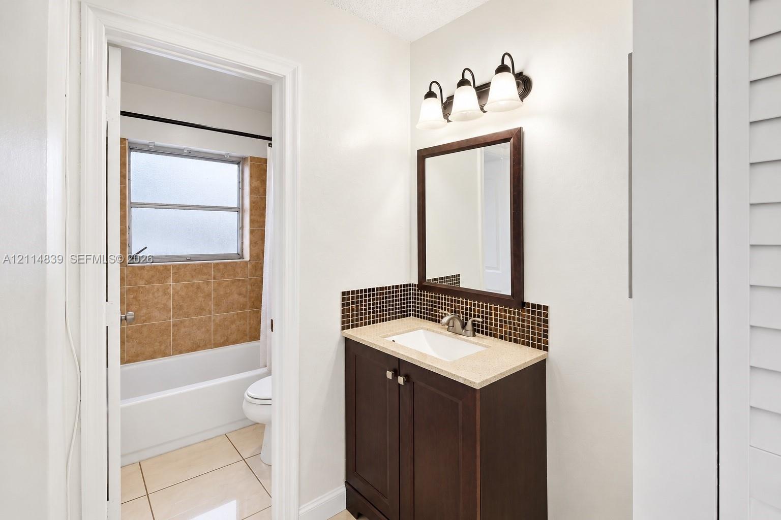 Bathroom featuring a dark wood-finish vanity with an integrated basin, stone-composite countertop, mosaic tile backsplash, and a three-light wall-mounted fixture
