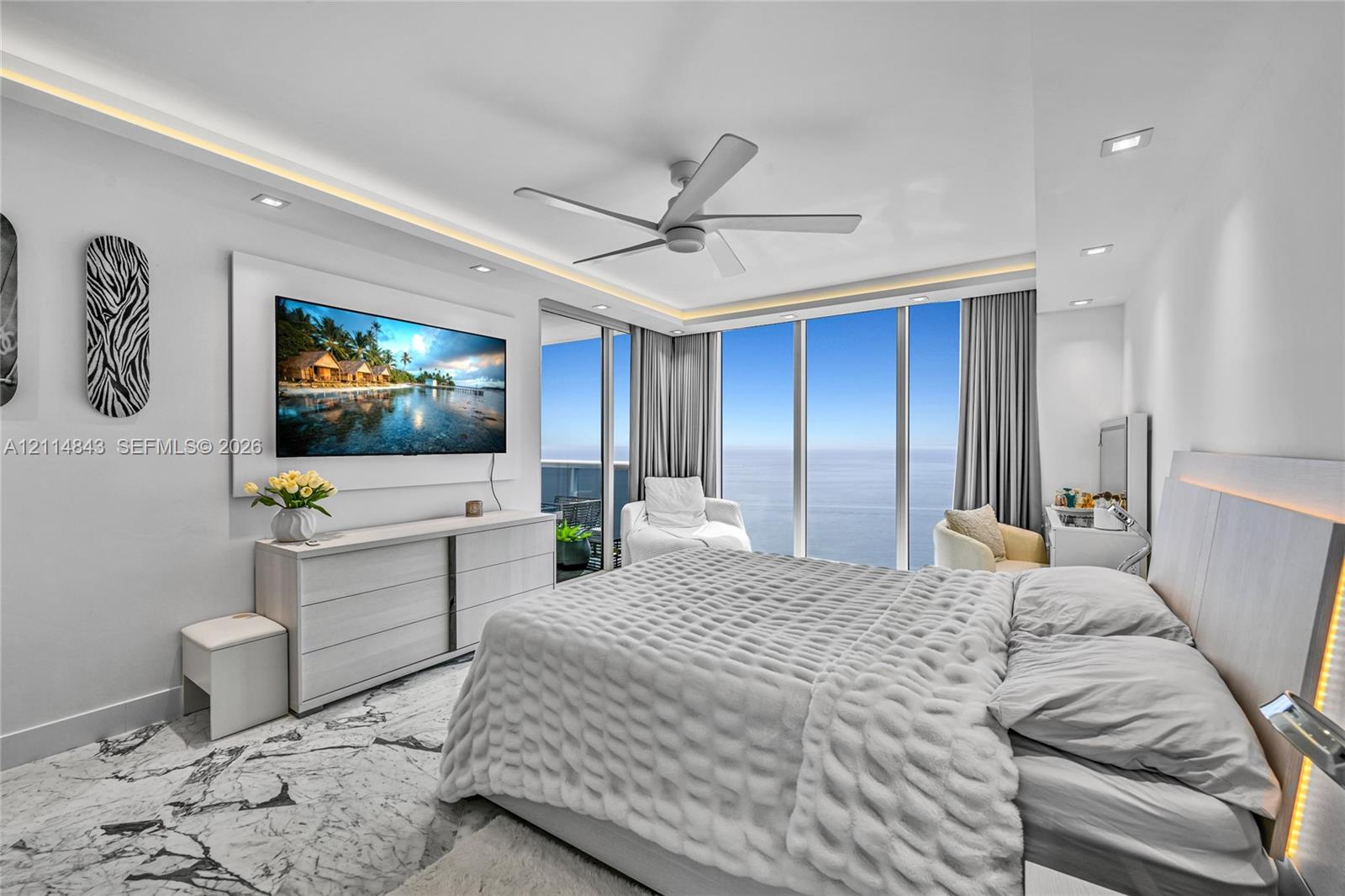 Floor-to-ceiling windows, light-toned stone-finish flooring, recessed LED cove lighting, modern ceiling fan, and built-in wall-mounted cabinetry