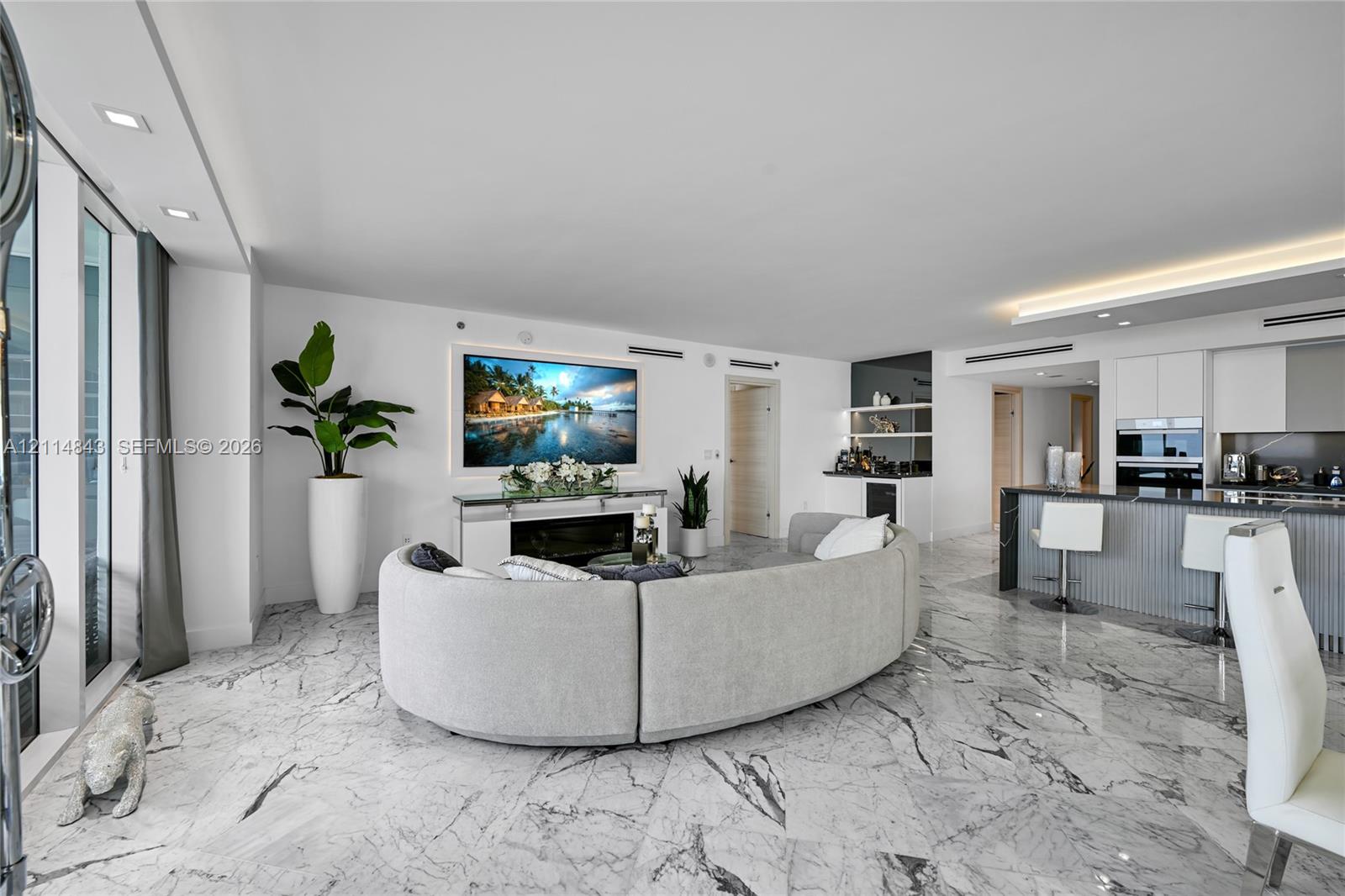 Open-concept living space featuring white stone-finish flooring, recessed lighting, built-in electric fireplace, recessed wet bar with shelving, and kitchen island with fluted cabinetry and integrated appliances
