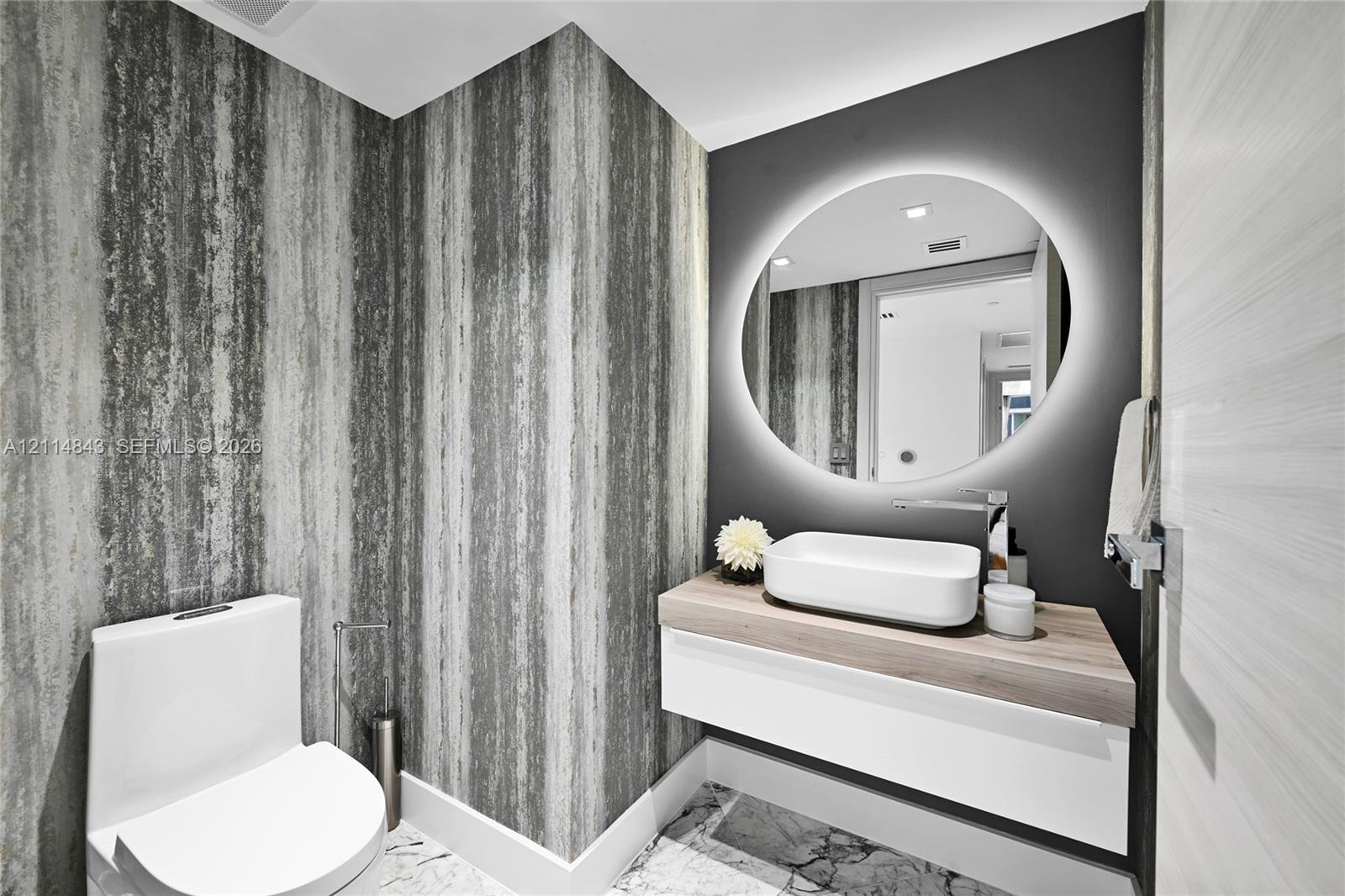 Floating vanity with wood-finish countertop, white vessel sink, polished chrome faucet, circular backlit mirror, and stone-patterned floor tile