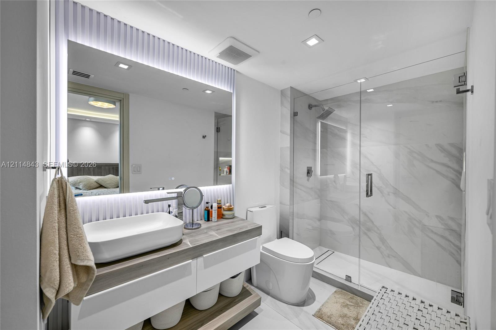 Floating vanity with a rectangular vessel sink and wood-finish countertop, full-height backlit mirror, walk-in glass shower enclosure with stone-finish tile surround, and recessed ceiling lighting