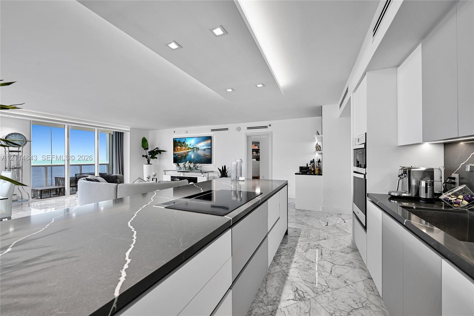 Modern kitchen featuring a large central island with veined stone countertop and integrated cooktop, handle-less cabinetry, built-in double ovens, and polished stone-finish flooring