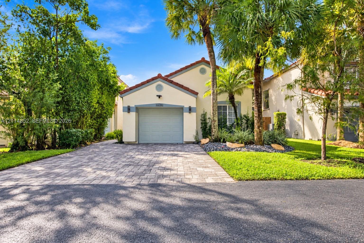 Stucco exterior featuring a tiled gable roof, paver driveway, single-car garage, and curated landscaping with mature palm trees
