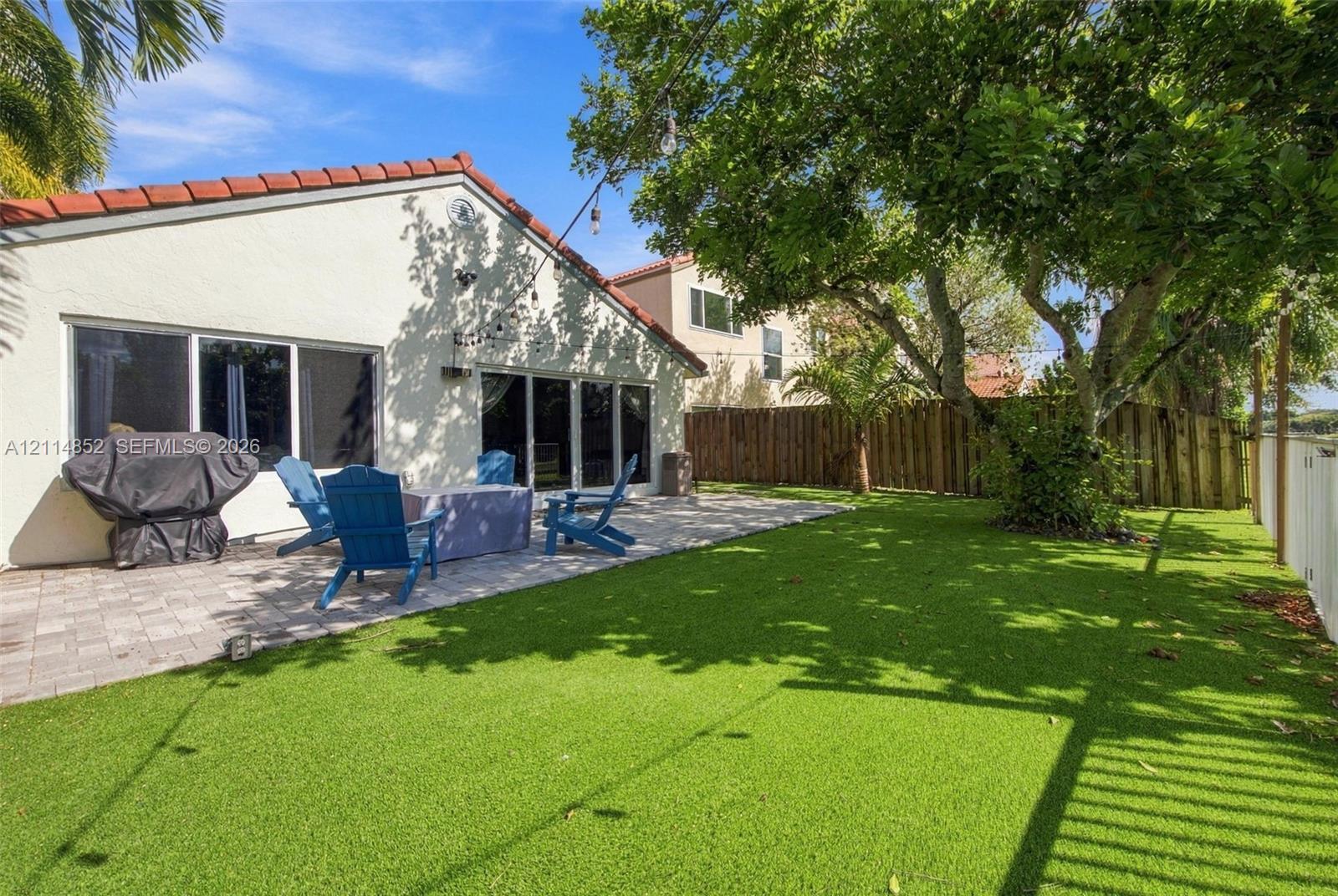 Backyard featuring a paved stone patio, synthetic lawn, and mature trees, enclosed by perimeter fencing
