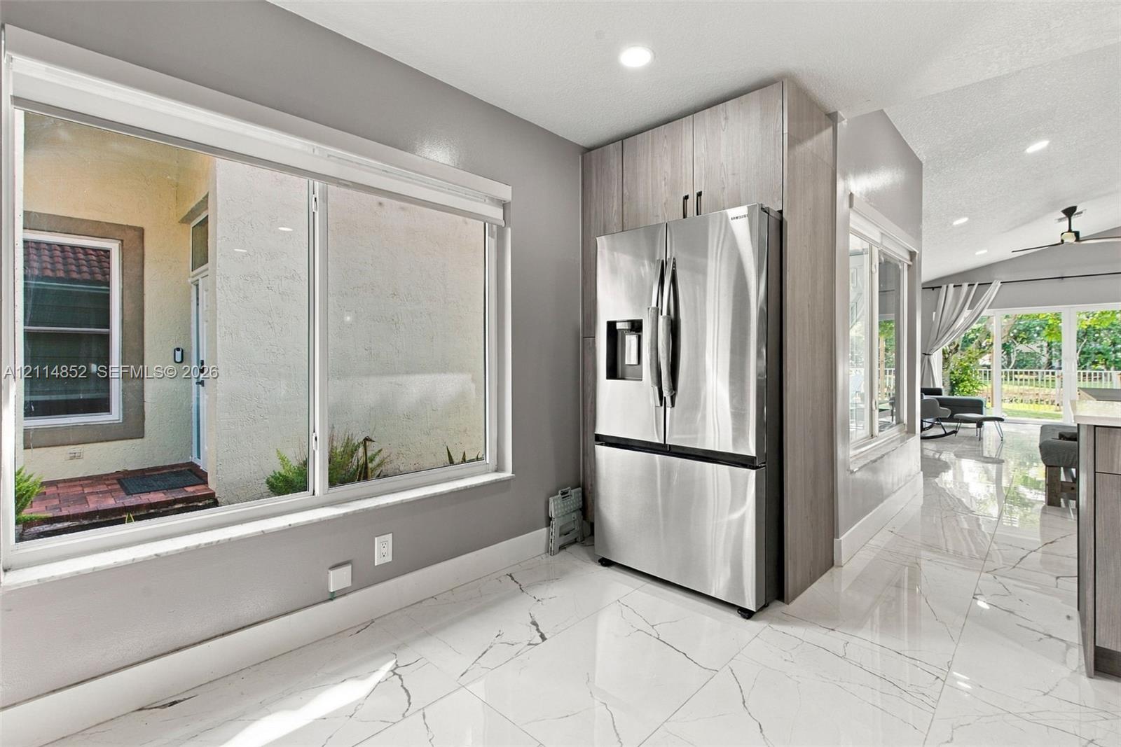 Large picture window, stainless steel French-door refrigerator, floor-to-ceiling wood-finish cabinetry, and polished stone-finish tile flooring with veining