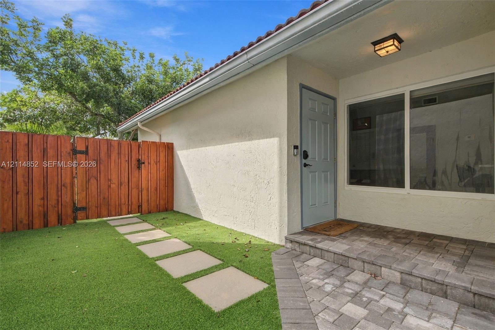 Exterior entryway featuring a stucco facade, a stone-paver porch, a wood-finish fence, and a stepped paver walkway set into synthetic turf