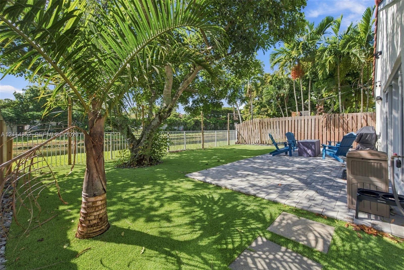 Private backyard featuring a paved stone patio, synthetic turf lawn, mature palm trees, and perimeter wood fencing