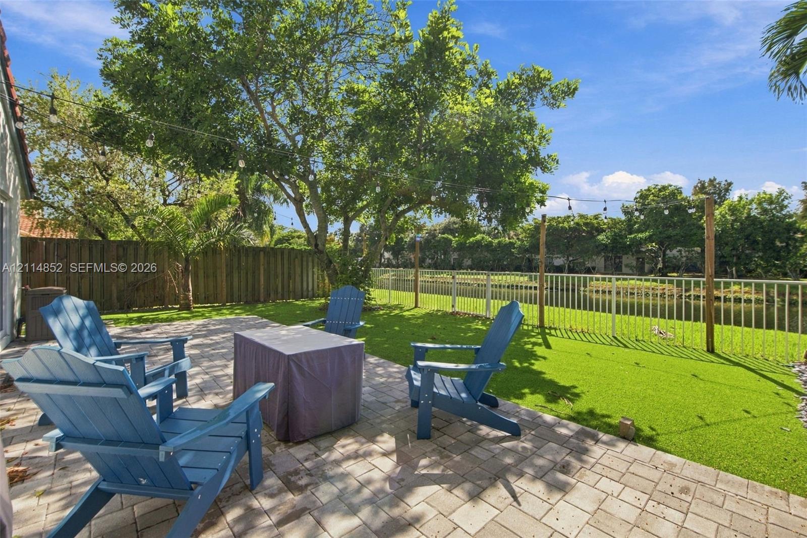 Backyard featuring a paved patio, perimeter wood fencing, water-facing white metal fencing, mature trees, and string lighting