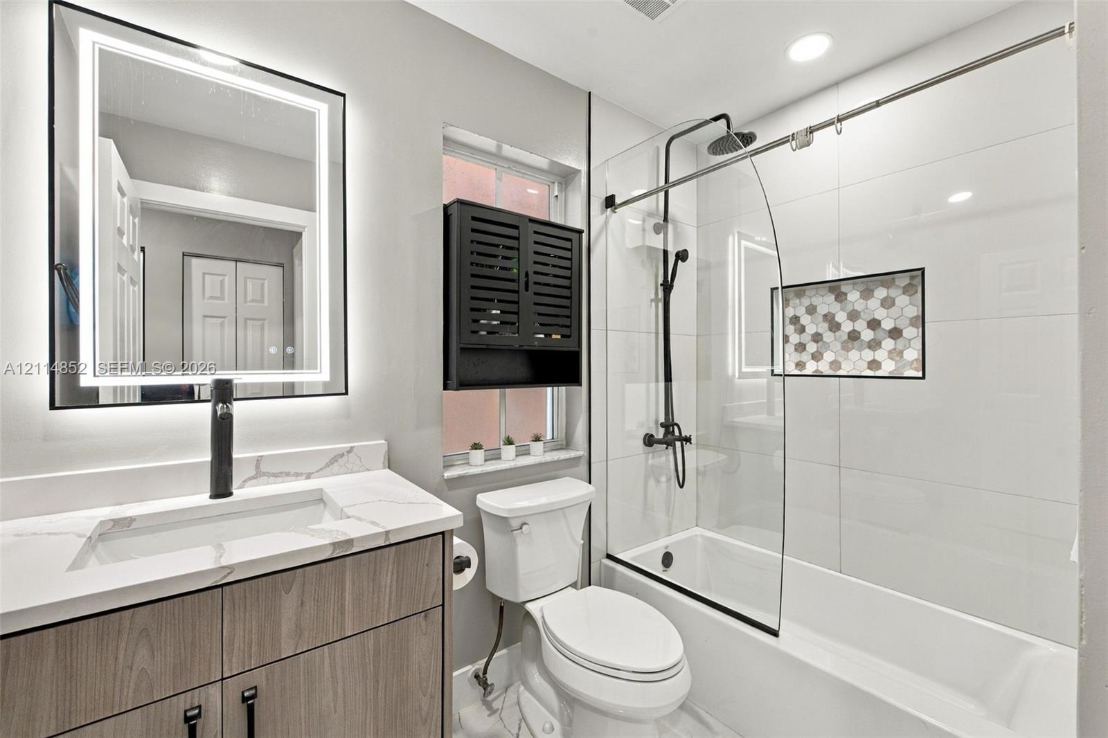 Bathroom featuring a wood-finish vanity with a stone-top integrated sink, matte black faucet, LED-backlit mirror, and a tub-shower combination with a glass panel and hexagon tile recessed niche