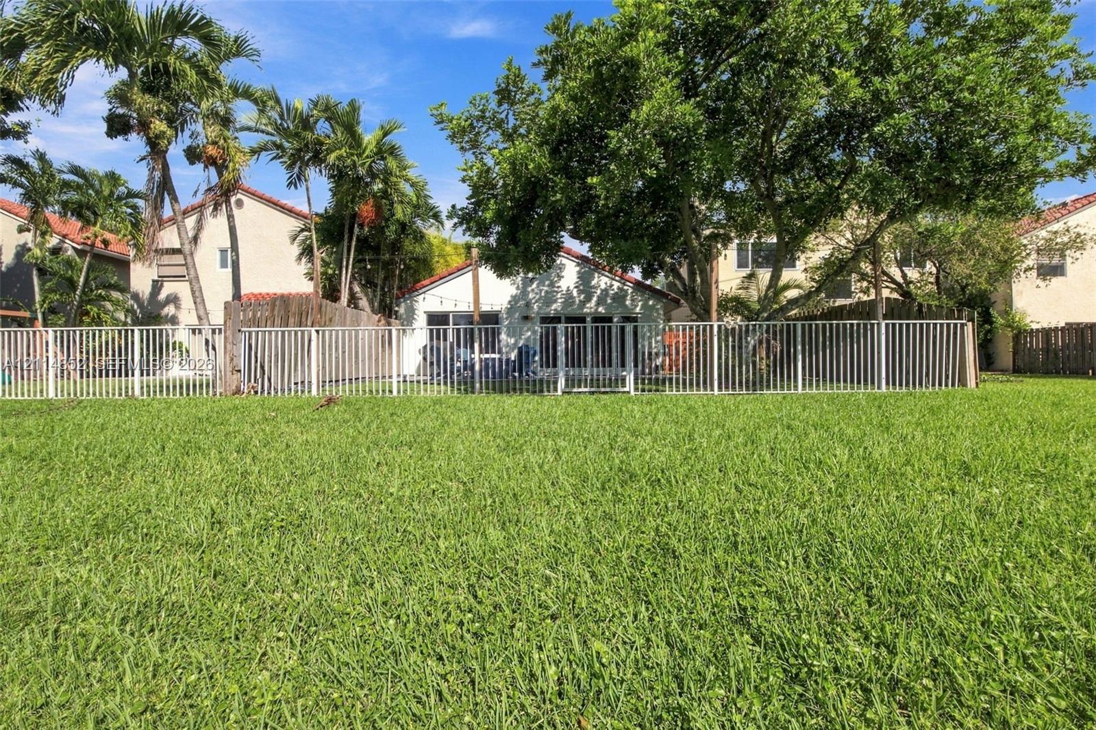 Expansive backyard featuring a manicured lawn, white metal fencing, mature trees, and palm trees