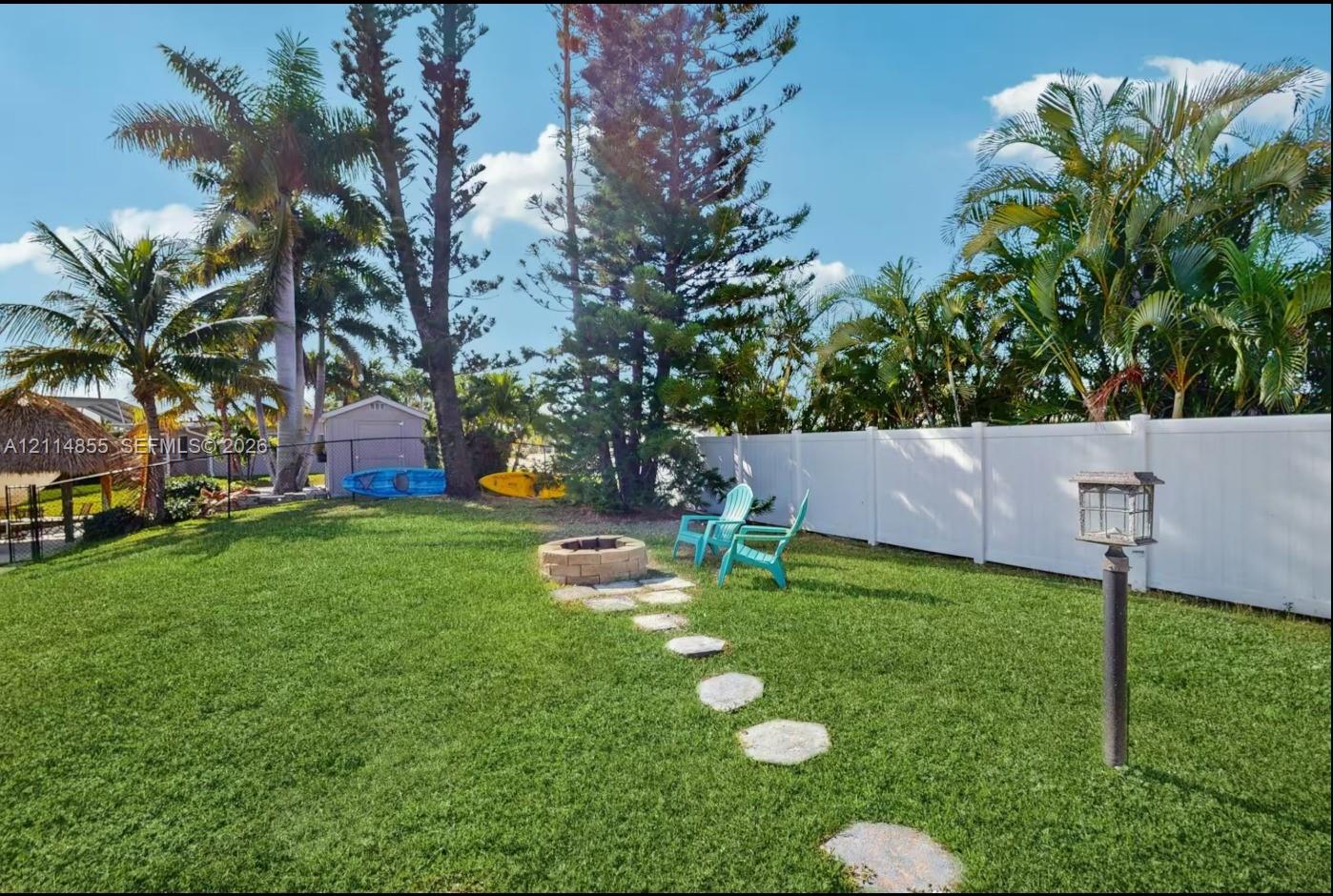 Fenced backyard featuring a manicured lawn, stone-paver path, stone fire pit, and mature palm trees