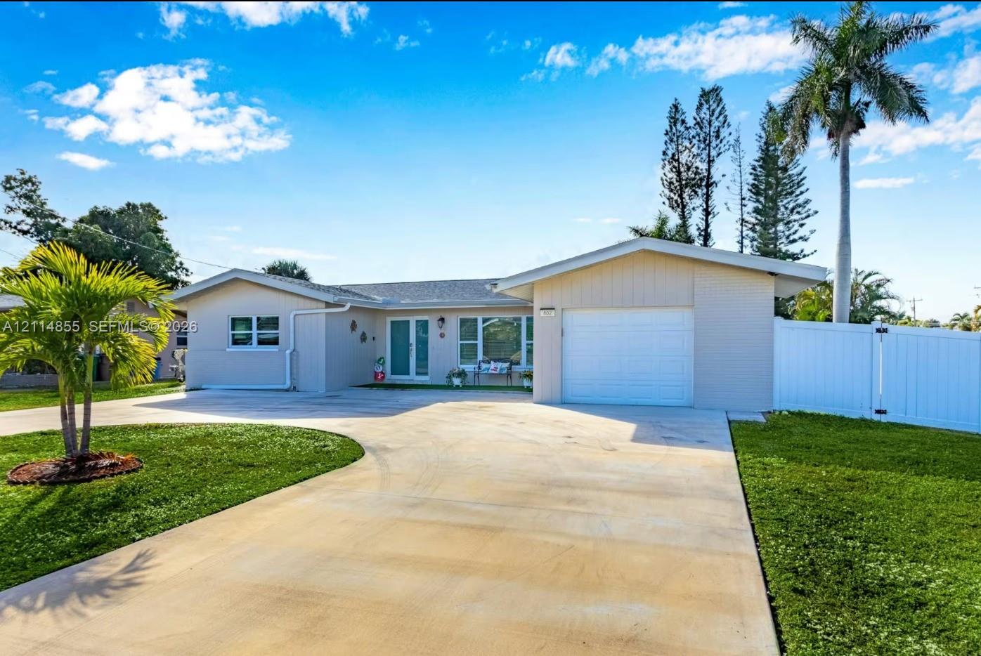 Single-story exterior featuring a concrete driveway, attached one-car garage, white vinyl fencing, and palm-accented landscaping