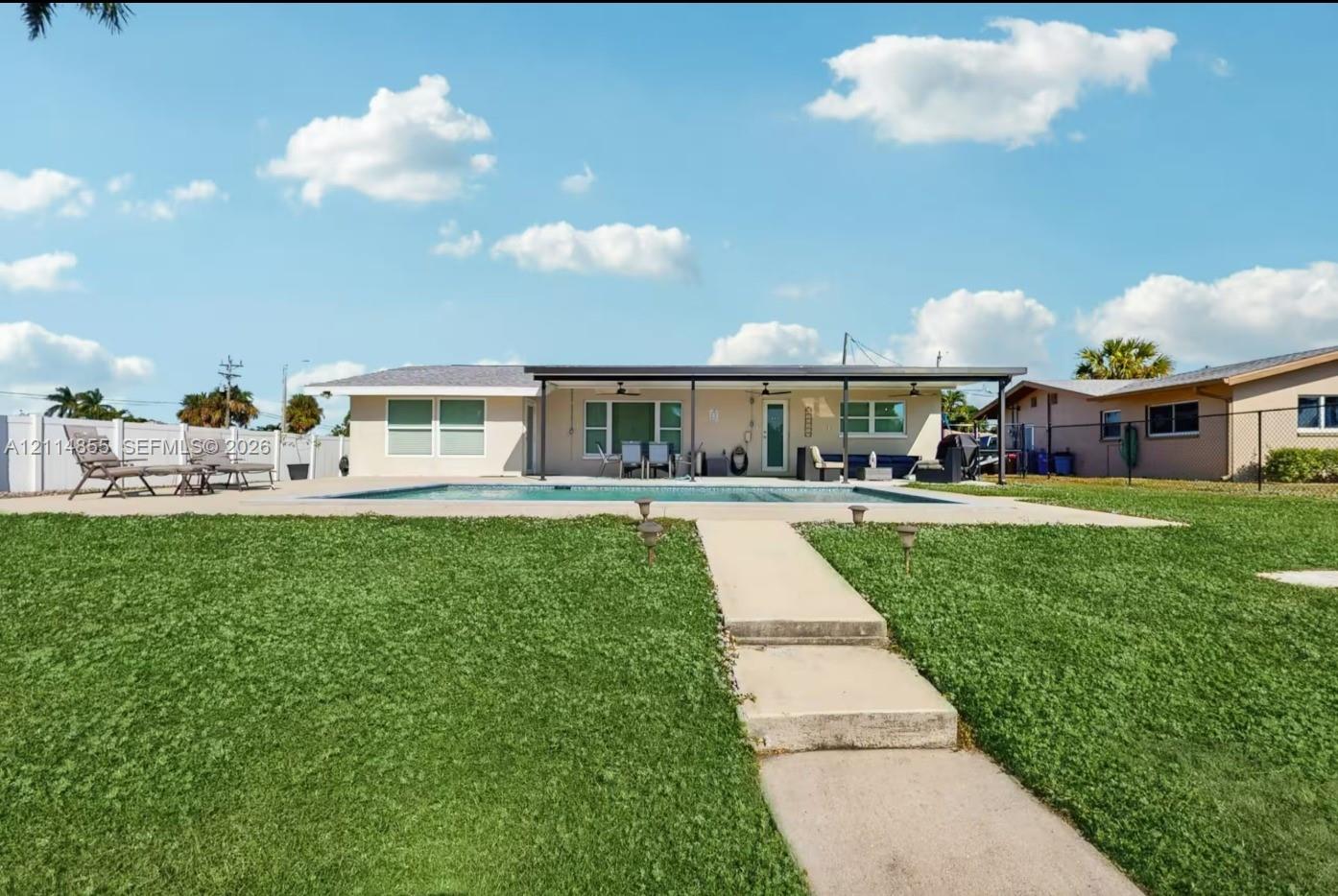 Exterior view featuring a rectangular swimming pool, concrete patio, expansive lawn, white vinyl fencing, and a covered porch with ceiling fans