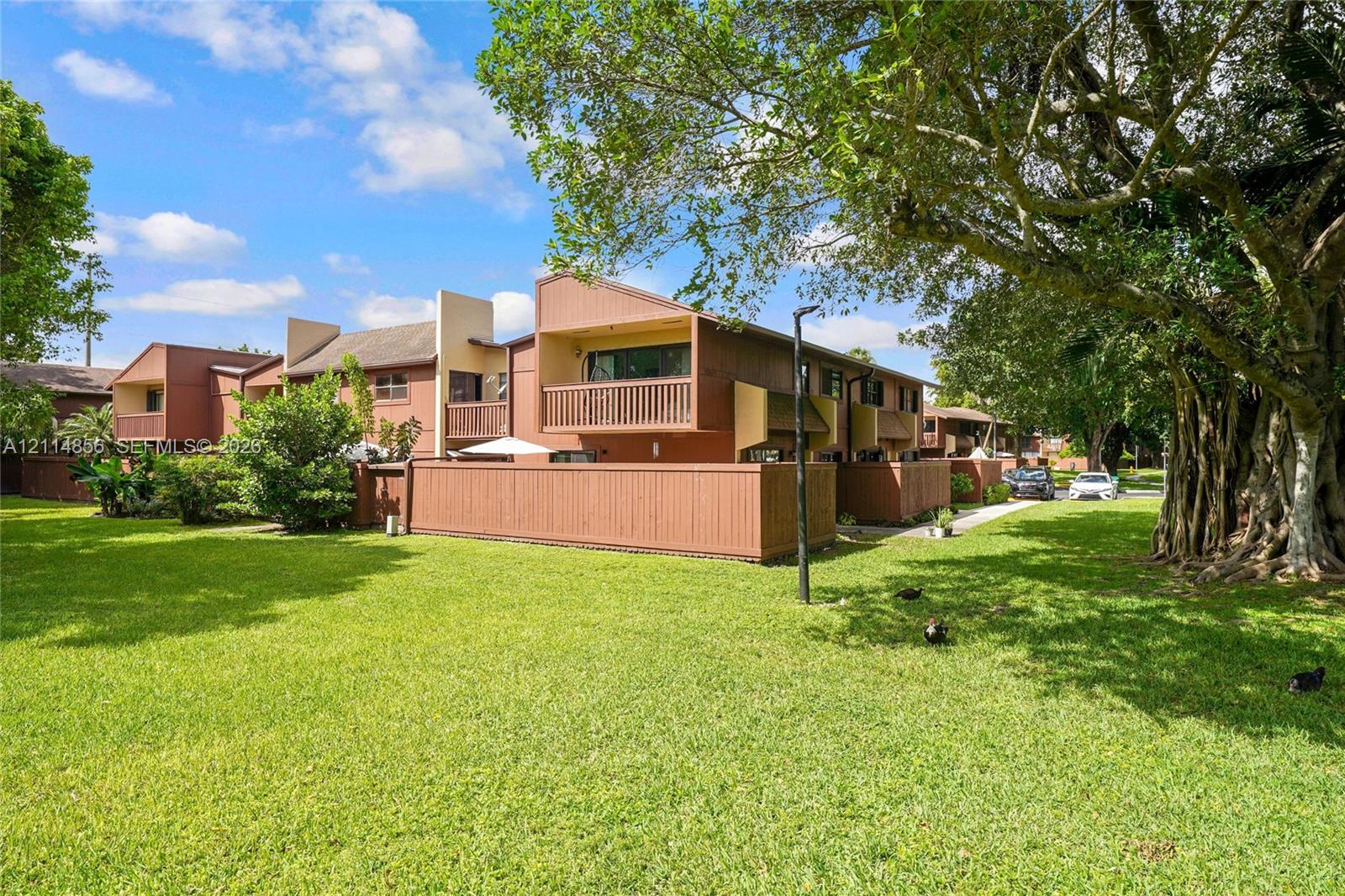 Multi-level residential exterior featuring horizontal wood siding, private balconies with wood railings, enclosed courtyard fencing, and expansive green space with mature trees