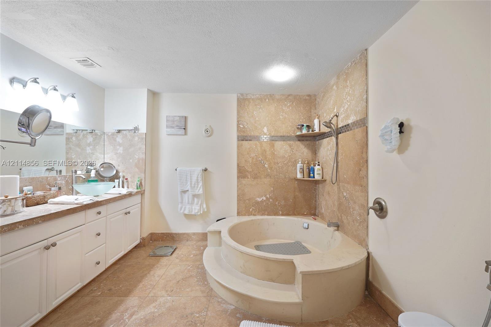 Bathroom featuring a stone-tile surround, circular soaking tub with a step-up entry, vanity with an integrated vessel sink, and stone-tile flooring