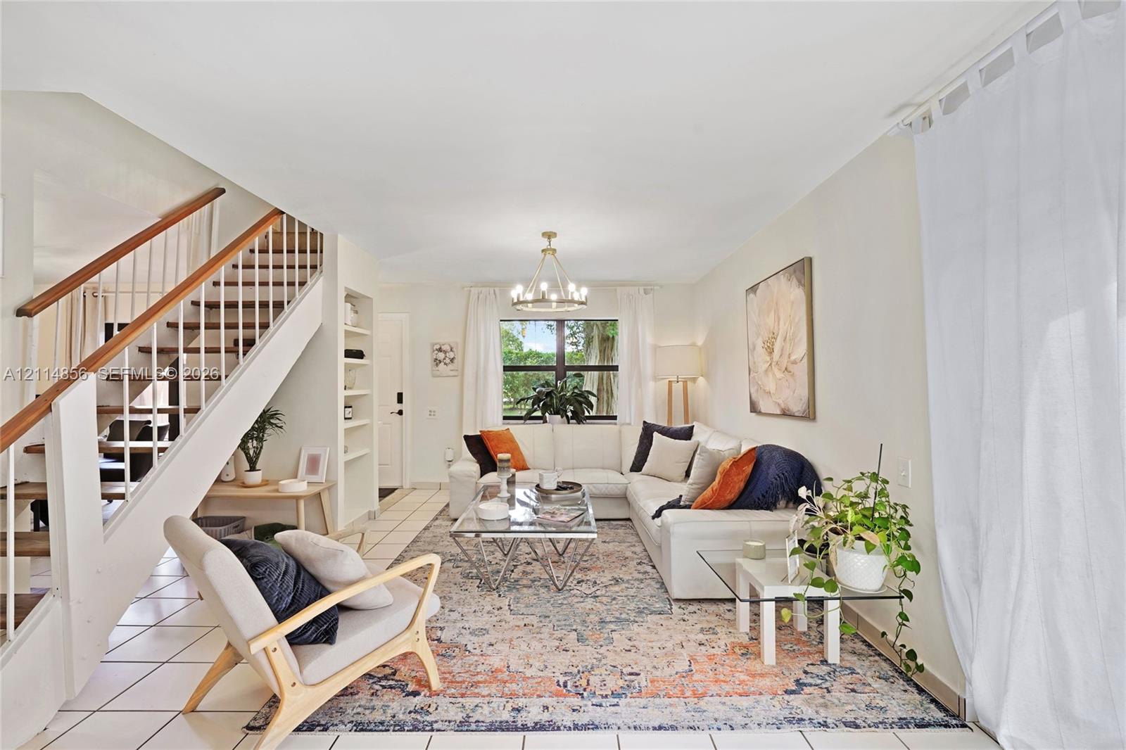 Open-concept living space featuring a central multi-arm chandelier, built-in recessed shelving, a staircase with wood-capped handrails, and tile flooring Open-concept living space featuring a central multi-arm chandelier, built-in recessed shelving, a staircase with wood-capped handrails, and tile flooring