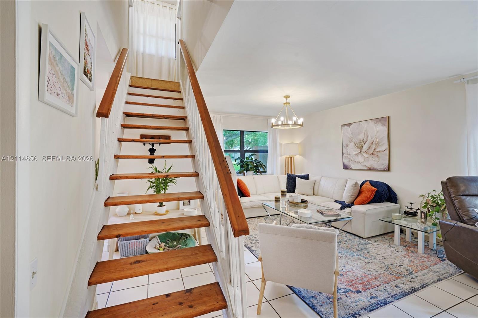 Open-concept living area featuring white tile flooring, wooden stair treads with white balusters, and a multi-arm modern chandelier