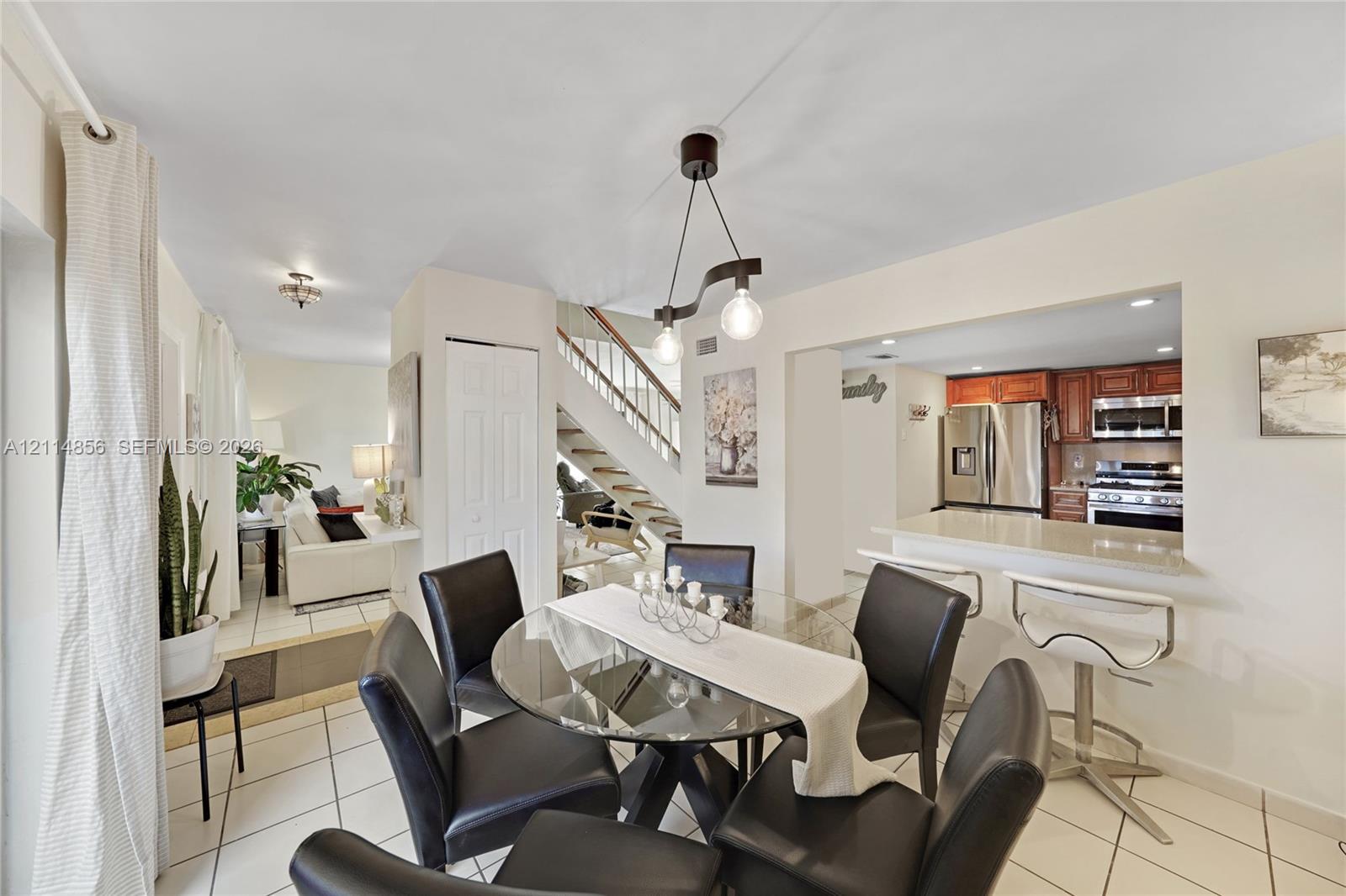 Open-concept interior featuring white ceramic tile flooring, a kitchen breakfast bar with light-toned solid surface countertops, wooden cabinetry, and a staircase with a wooden handrail Open-concept interior featuring white ceramic tile flooring, a kitchen breakfast bar with light-toned solid surface countertops, wooden cabinetry, and a staircase with a wooden handrail