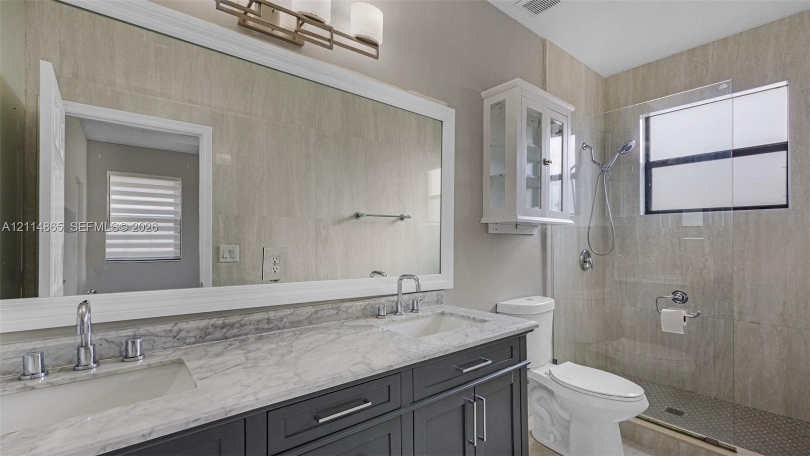 Dual-sink vanity with stone countertop and cabinetry, large framed mirror, wall-mounted glass cabinetry, and walk-in shower with glass enclosure